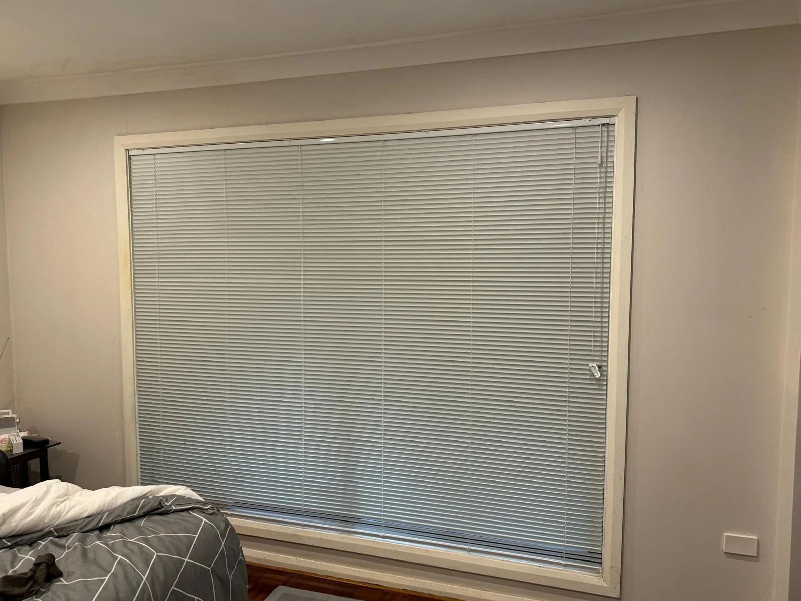 A bedroom with a large window covered by closed white blinds, beige walls, part of a bed with a gray and white geometric patterned blanket, a small nightstand with a clock and other items, and a light switch on the wall.