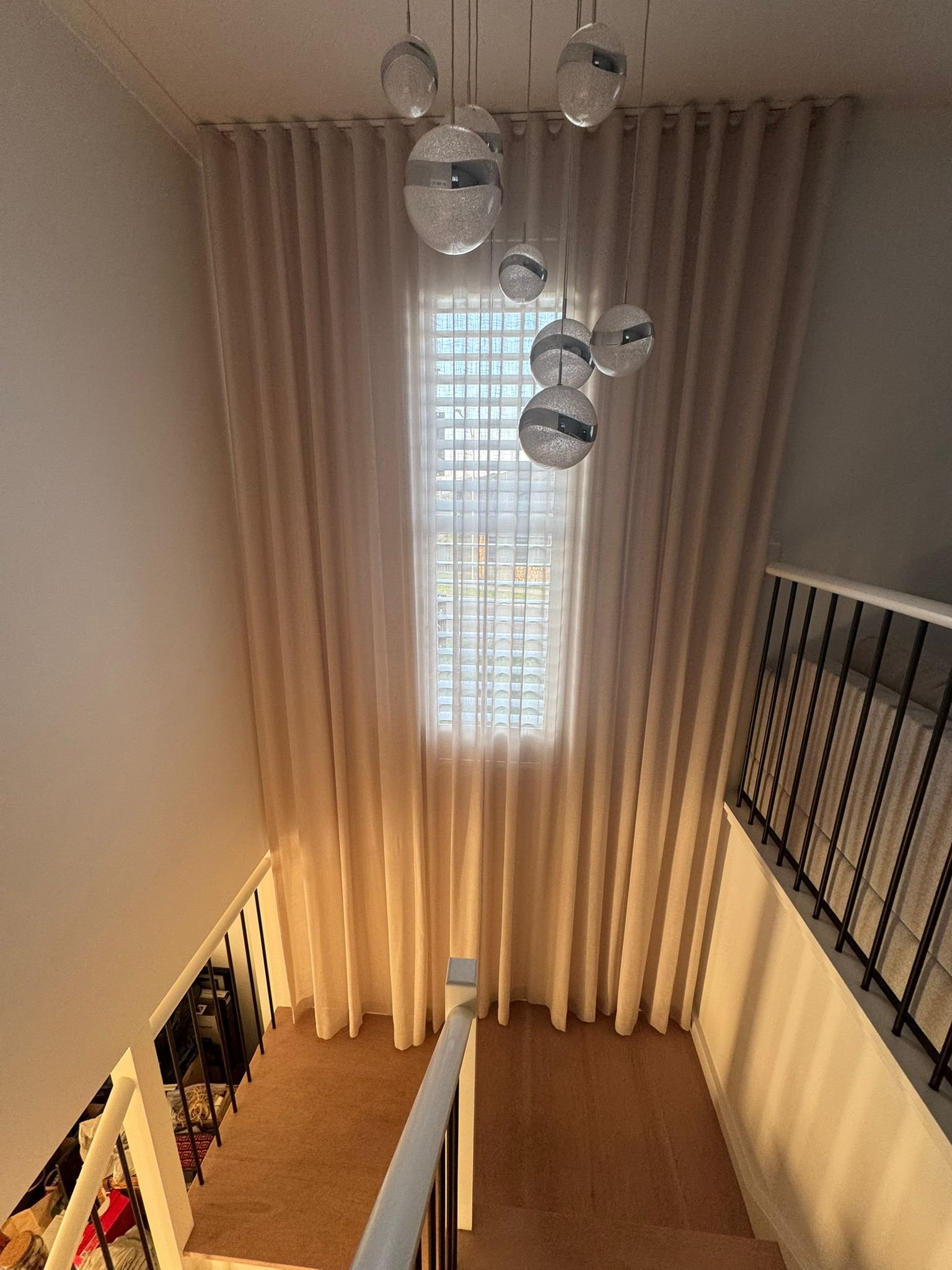 A staircase landing with a beige curtain hanging in front of window, a modern chandelier with spherical glass shades, and a black metal railing along the stairs.