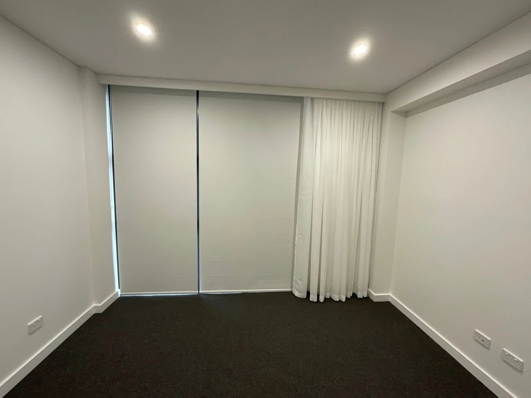 An empty room with white walls, a dark carpeted floor, a large sliding door closet with white curtains on the right, and a white ceiling with two recessed lights.