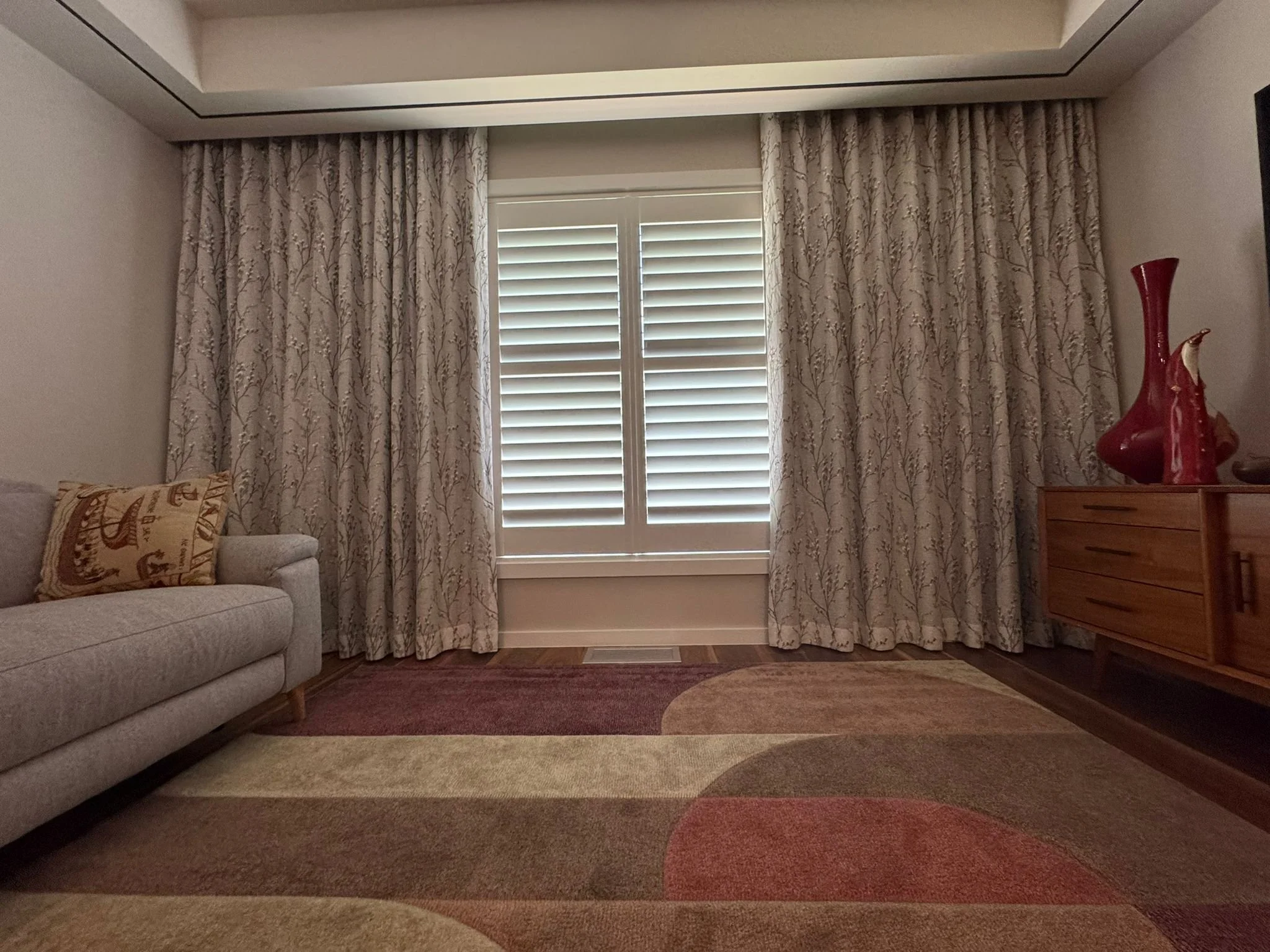 Living room with large window covered by patterned curtains, beige sofa with decorative pillow, wooden dresser with red vase, area rug, and hardwood floor.