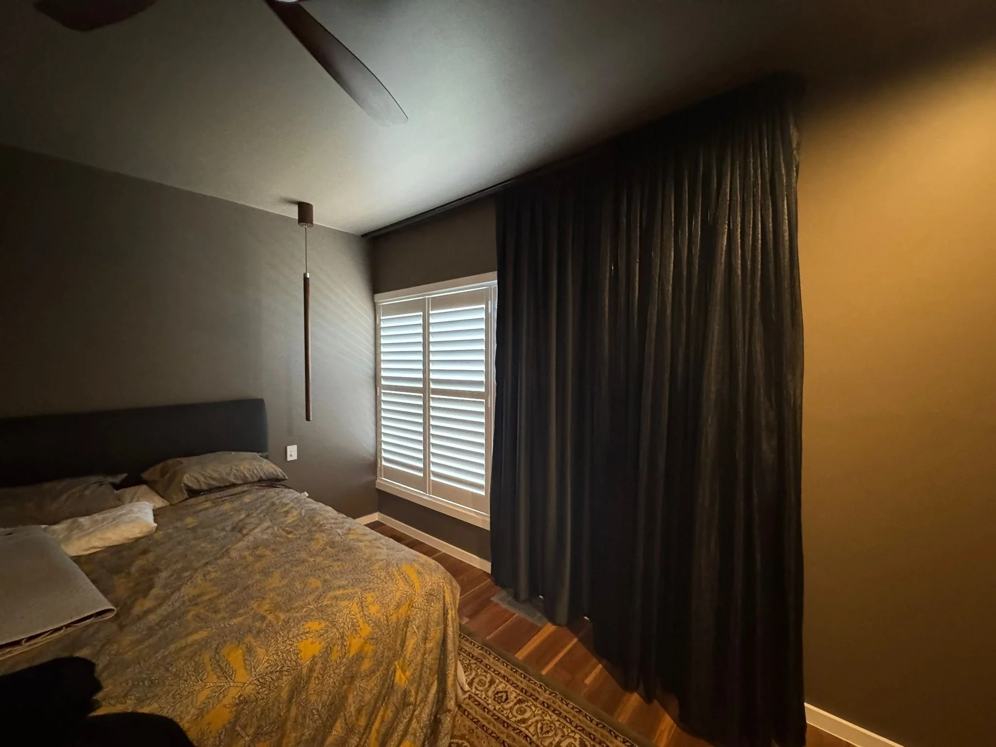 A bedroom with a bed, a window with shutters, dark curtains, a ceiling fan, and a pendant light.