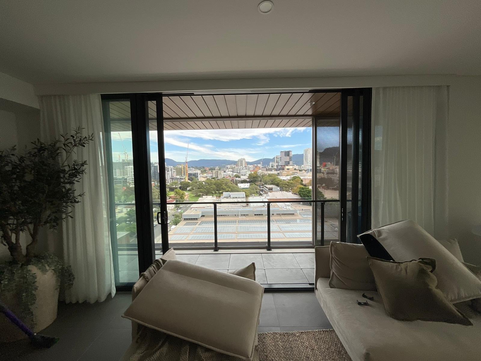 Living room with large glass sliding doors opening to a balcony overlooking a cityscape with buildings, trees, and mountains in the distance.