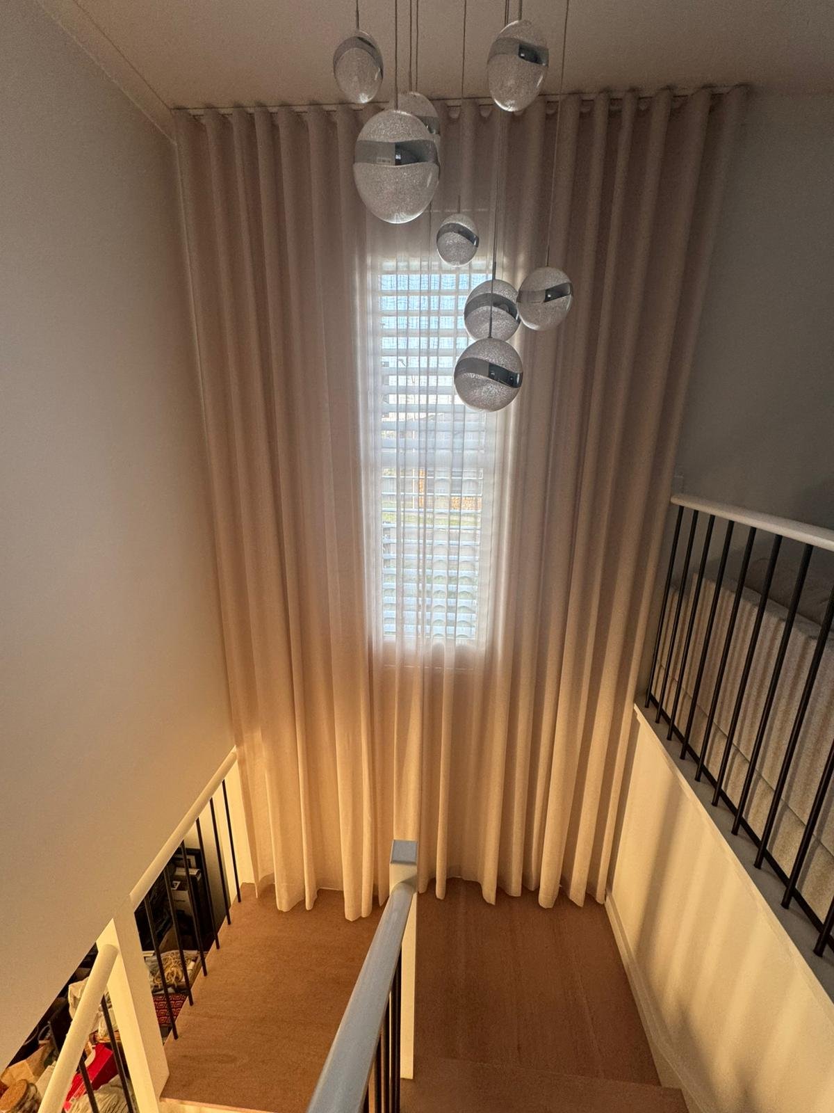 View from the top of a staircase looking up at a ceiling with hanging pendant lights and beige curtains covering a window, with railings on either side.