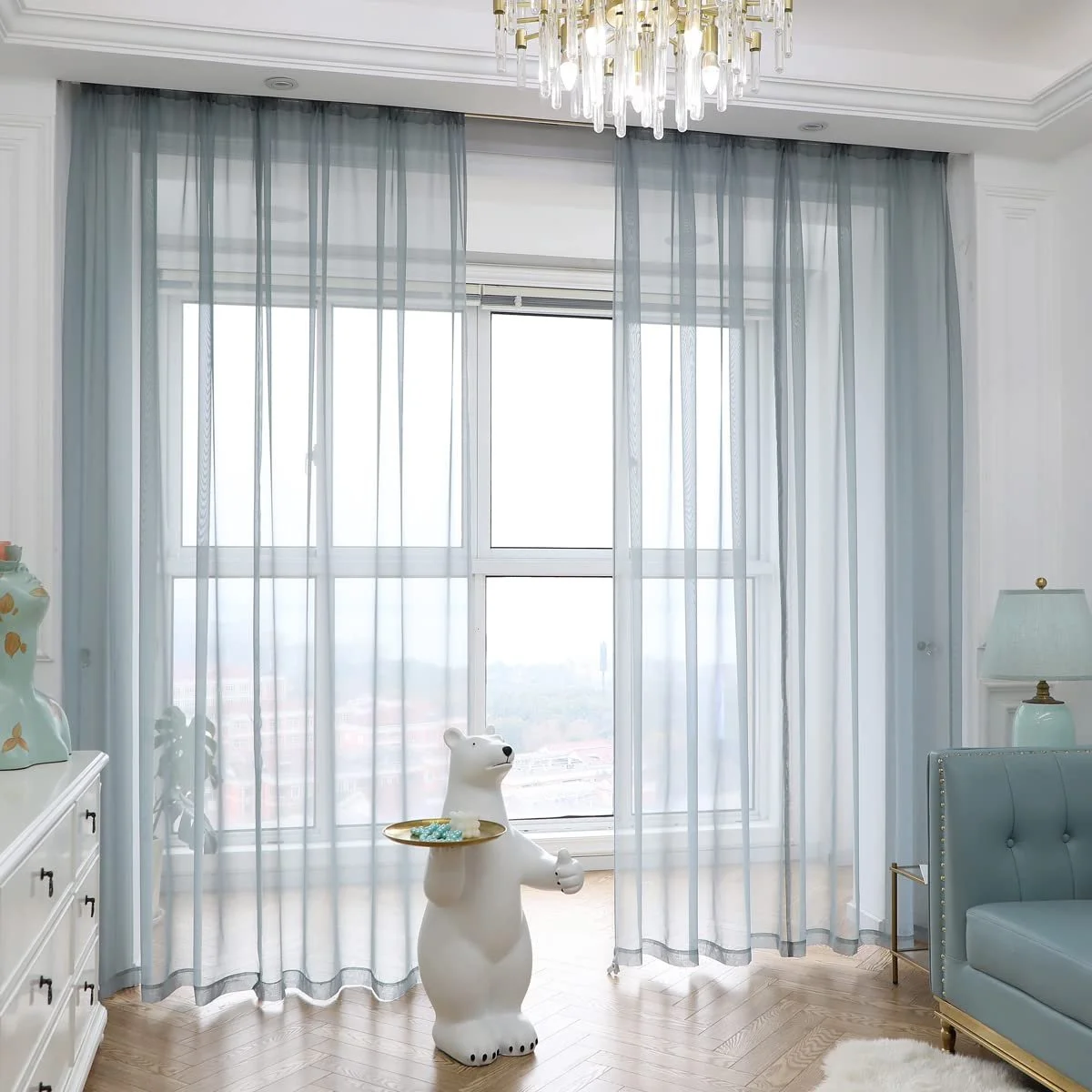 Living room with sheer curtains on large window, polar bear-shaped lamp holding a tray, white dresser with ceramic vase, pastel blue armchair, and table lamp, light-colored wooden floor.