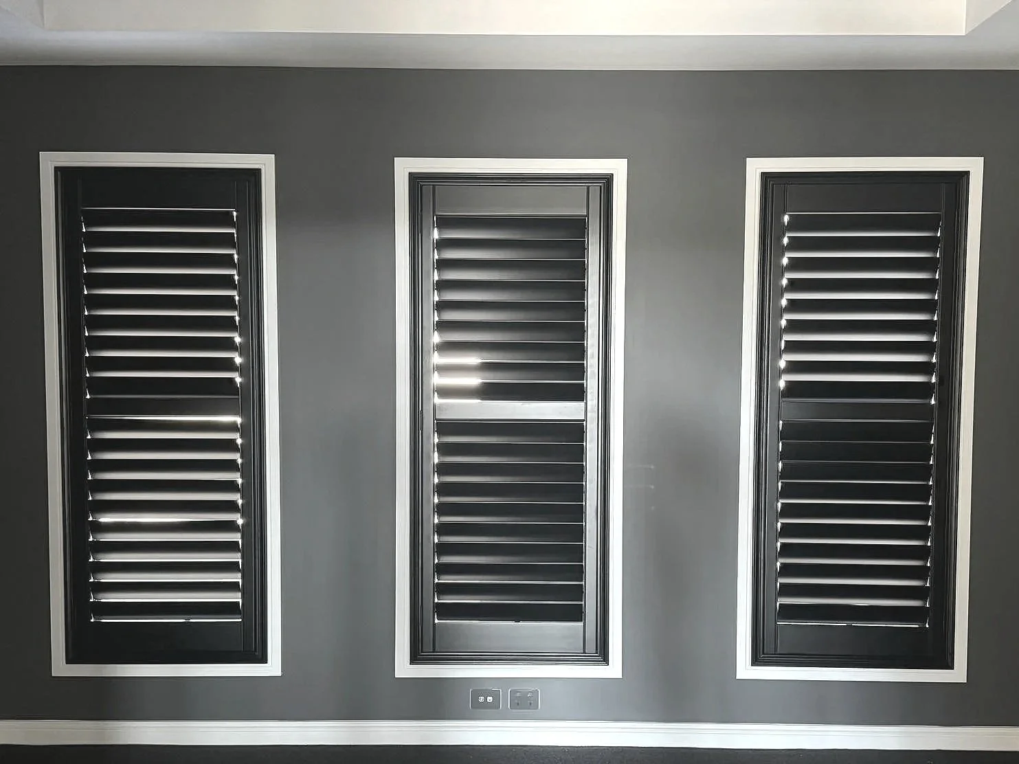 Three rectangular black window shutters with horizontal slats, framed in white, mounted on a dark gray wall. The shutters are closed, with light peeking through the gaps between the slats.