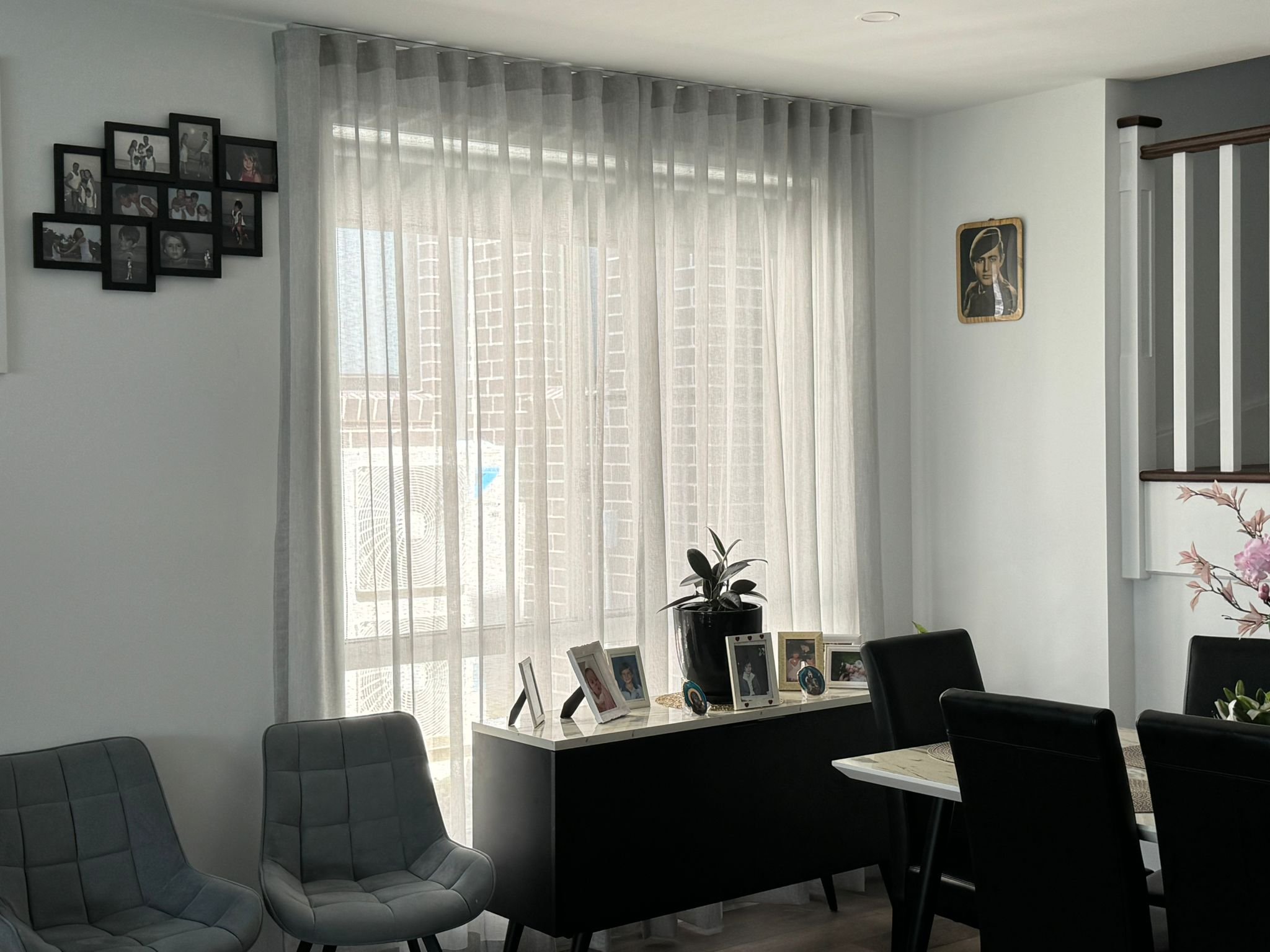 A bright dining or living room with sheer white curtains covering large windows. The room has black and gray chairs around a dining table, with photo frames and a potted plant on a white sideboard beneath the windows. There are framed pictures on the