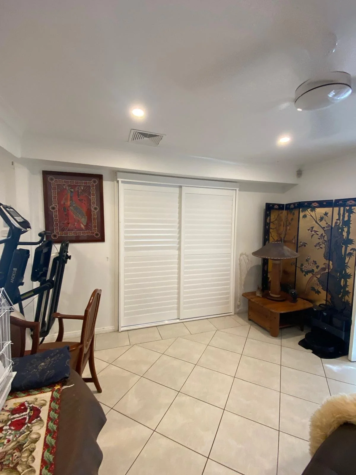 Living room corner with white louvered closet doors, a decorative folding screen with trees, wooden furniture, a lamp, a framed artwork, exercise equipment, and tiled flooring.