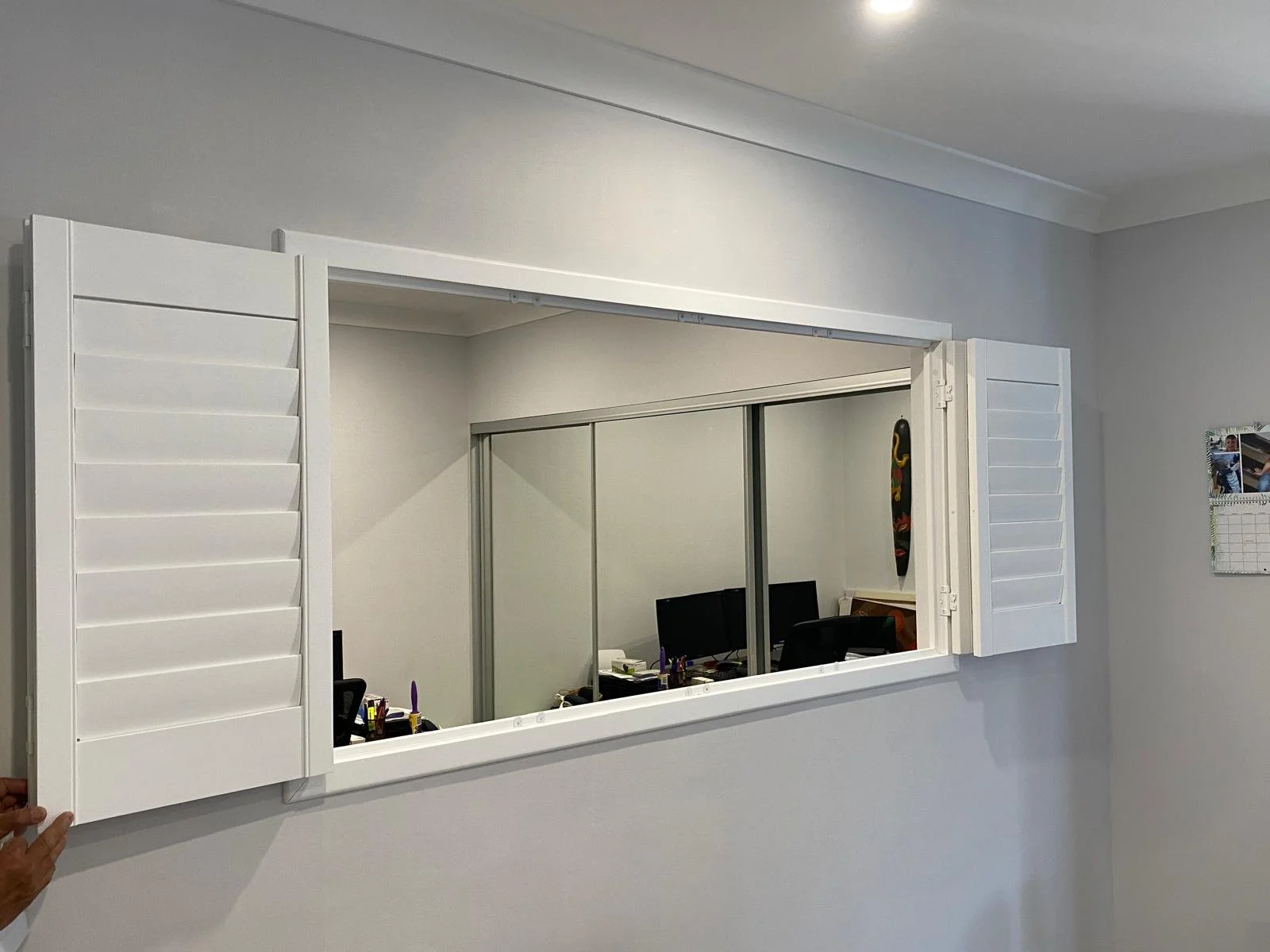 A wall mirror with white window shutters as a frame, reflecting a home office with two computer monitors, a desk, and some office supplies.