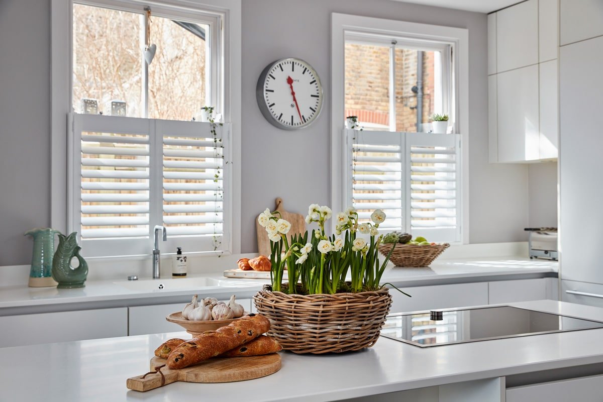 Bright, modern kitchen with white cabinets, a white countertop, and two windows with white shutters. A large basket of white flowers sits on the counter, along with garlic, breadsticks, and a tray of croissants. A wall clock is mounted between the windows, showing the time as 3:20.