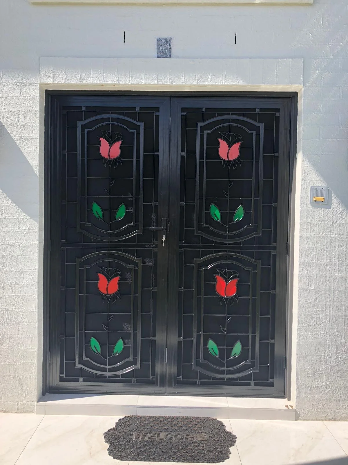 Black metal double door with stained glass roses in red and green, set in white brick wall with a welcome mat in front.
