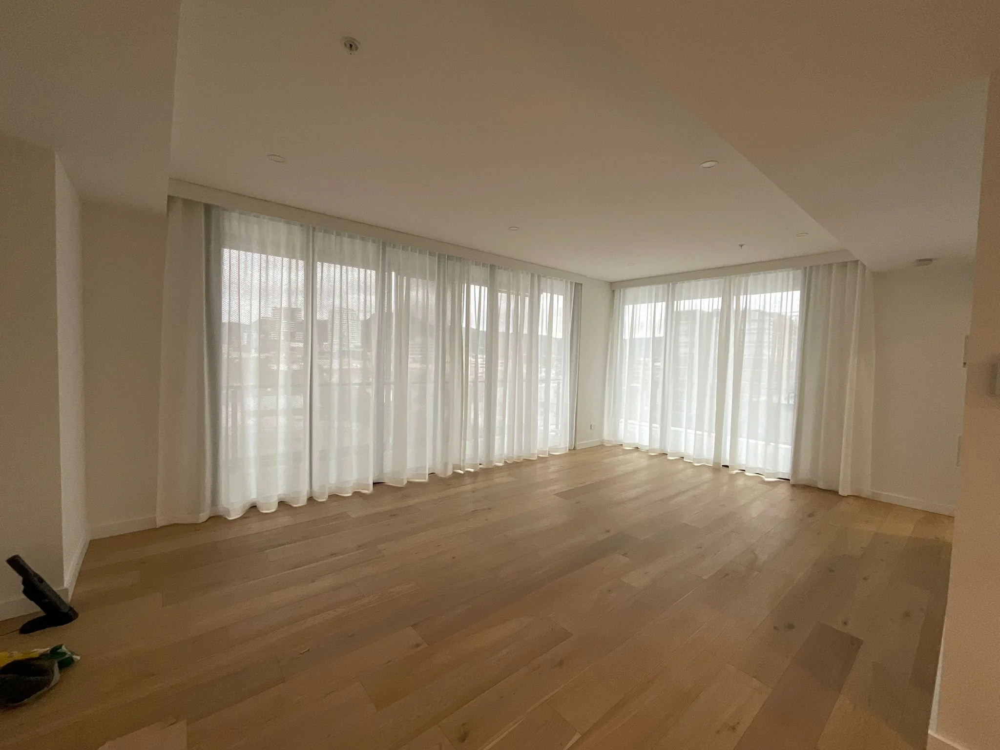 A bright room with large windows covered with sheer white curtains, light-colored hardwood floors, and a small black exercise bike in the left corner.