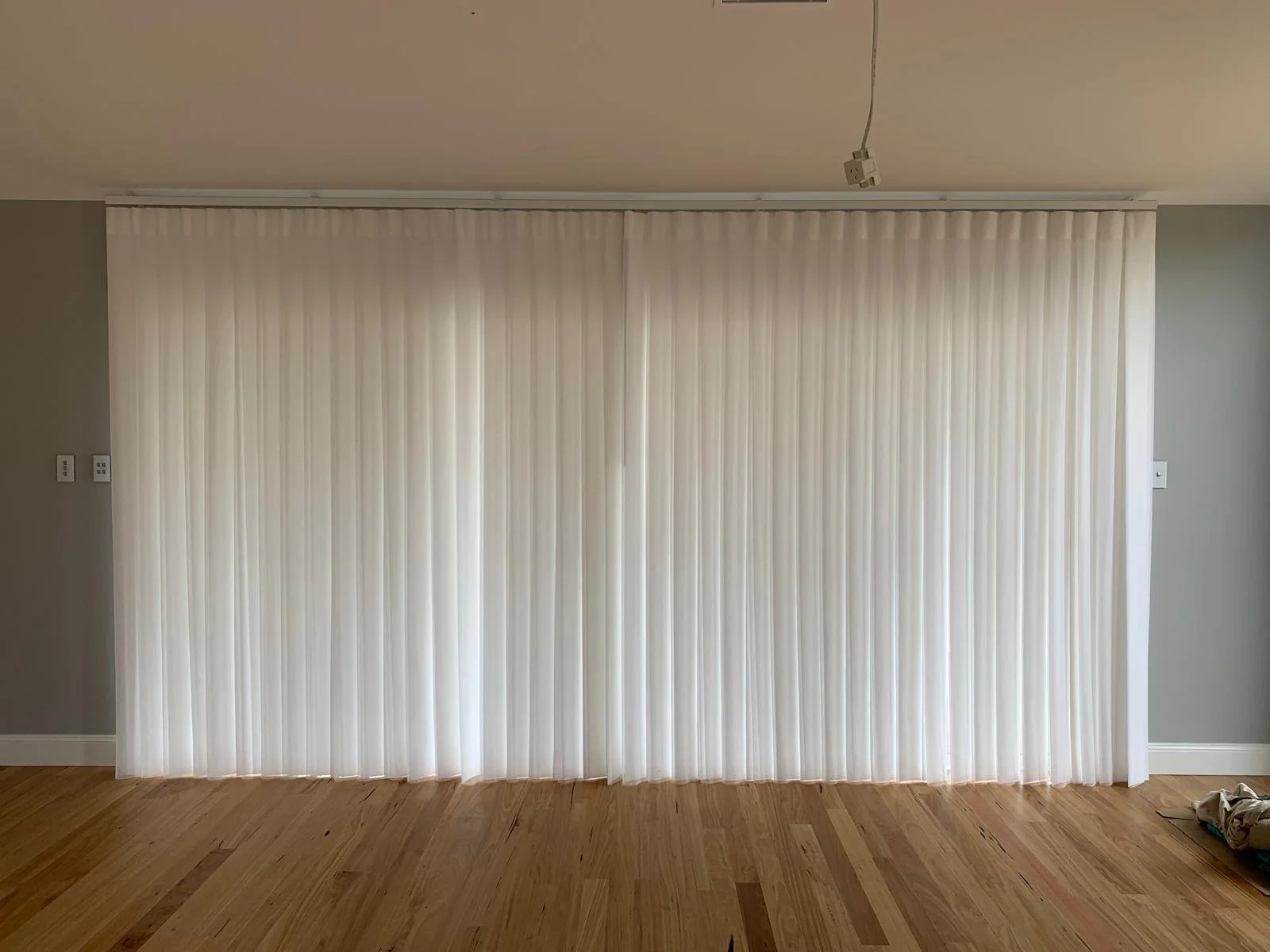 White vertical blinds covering a large window or sliding door in a room with light gray walls and wooden floors.