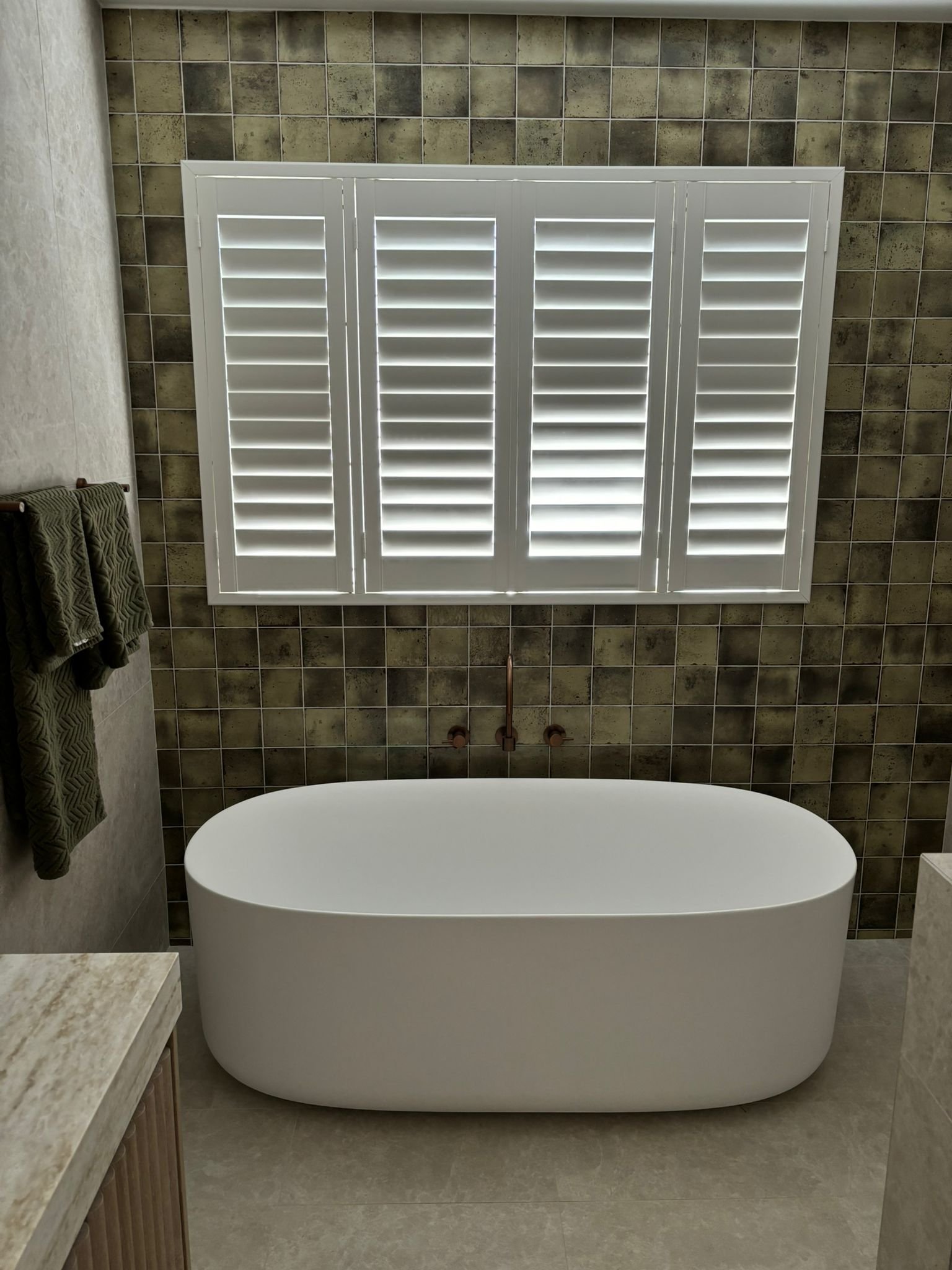 A modern bathroom with a large freestanding oval white bathtub, a window with white shutters, a tiled wall with earthy tones, and a stone countertop on the left.