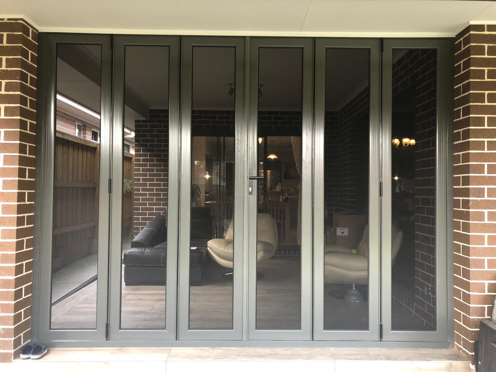 View of a modern enclosed porch with large glass sliding doors, black brick interior walls, and furniture including a black couch and beige chairs.