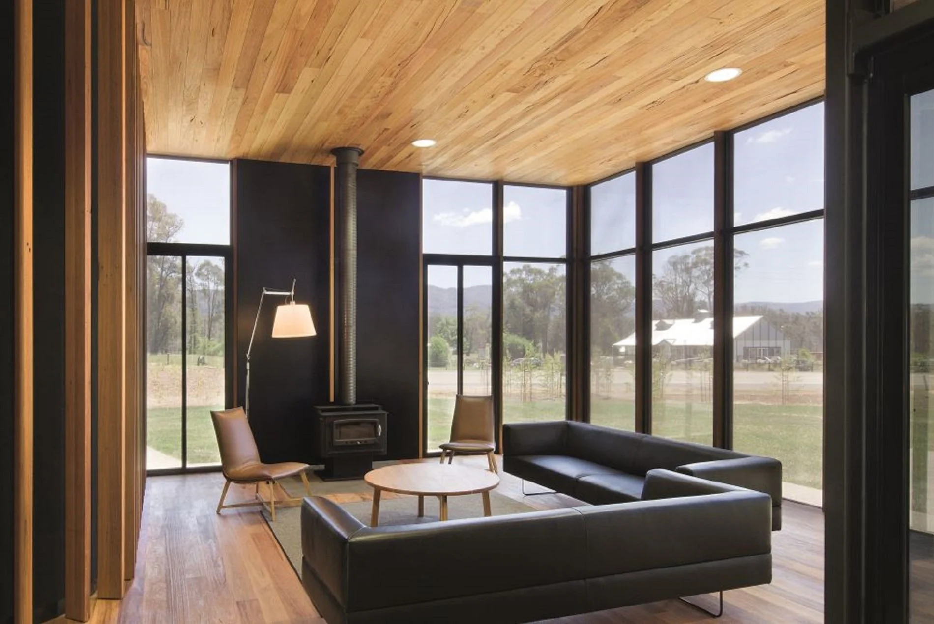 Modern living room with floor-to-ceiling windows, wooden ceiling, black wall with a wood stove pipe, black sofa, round wooden coffee table, and chairs, with a view of trees and a house outside.