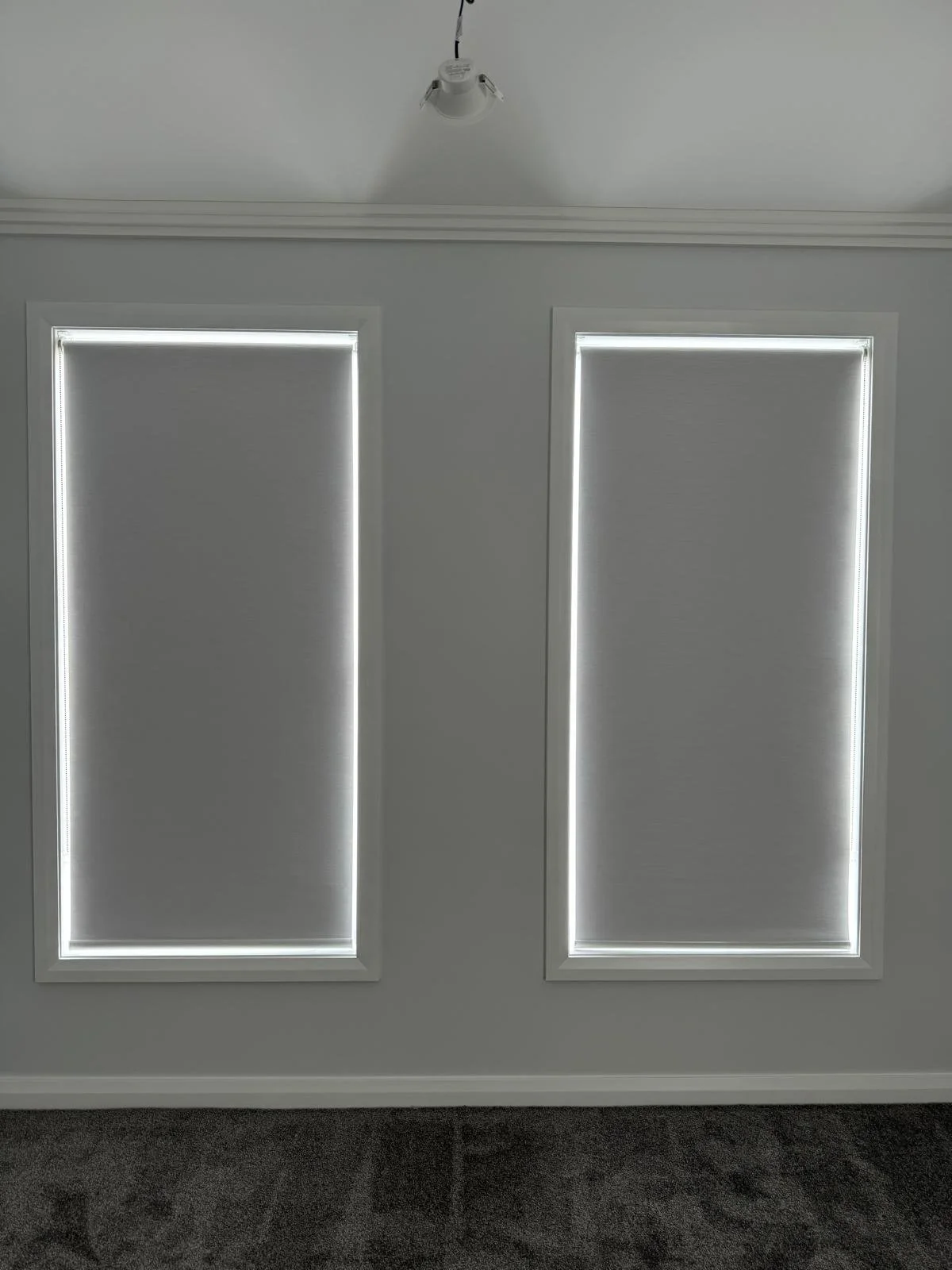 A minimalist interior room with two tall, narrow windows covered with white blinds and framed with white trim, and a ceiling mounted light fixture. The wall is painted white and the floor appears to be dark-colored.