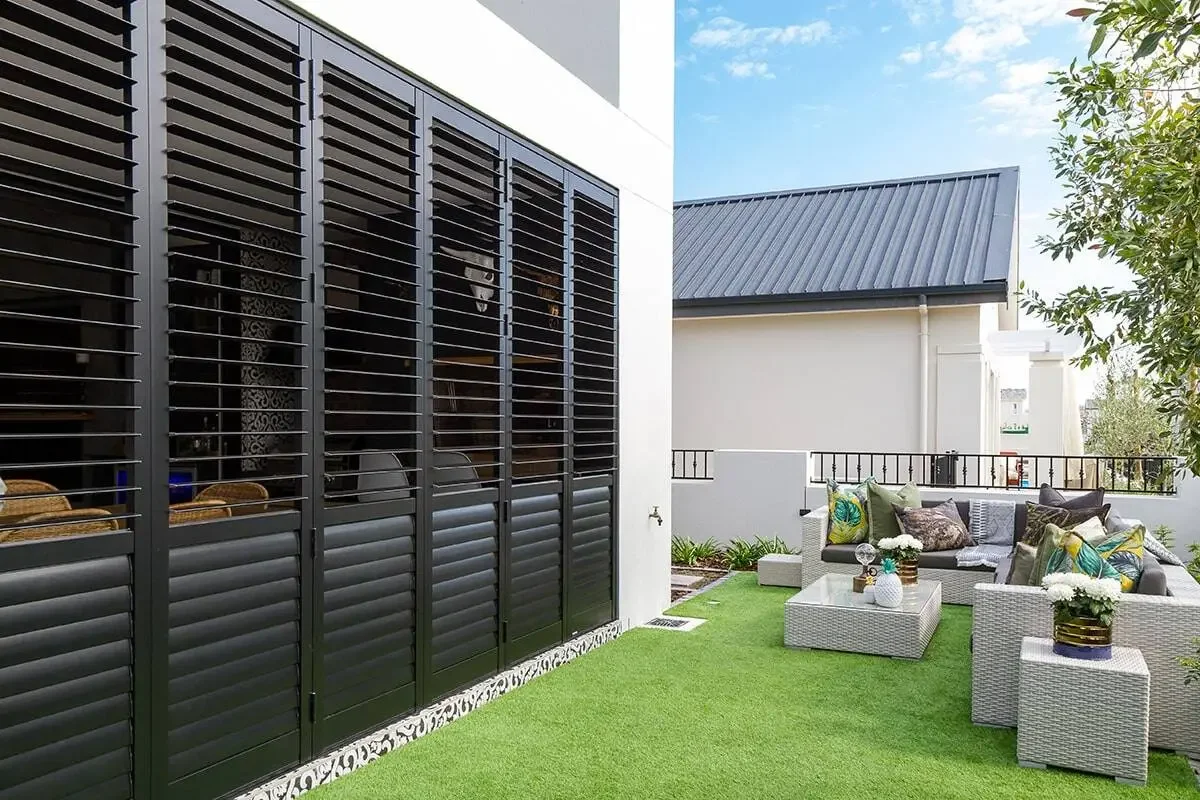 Outdoor patio area with gray wicker furniture, colorful cushions, and potted plants, surrounded by green artificial grass and white walls.
