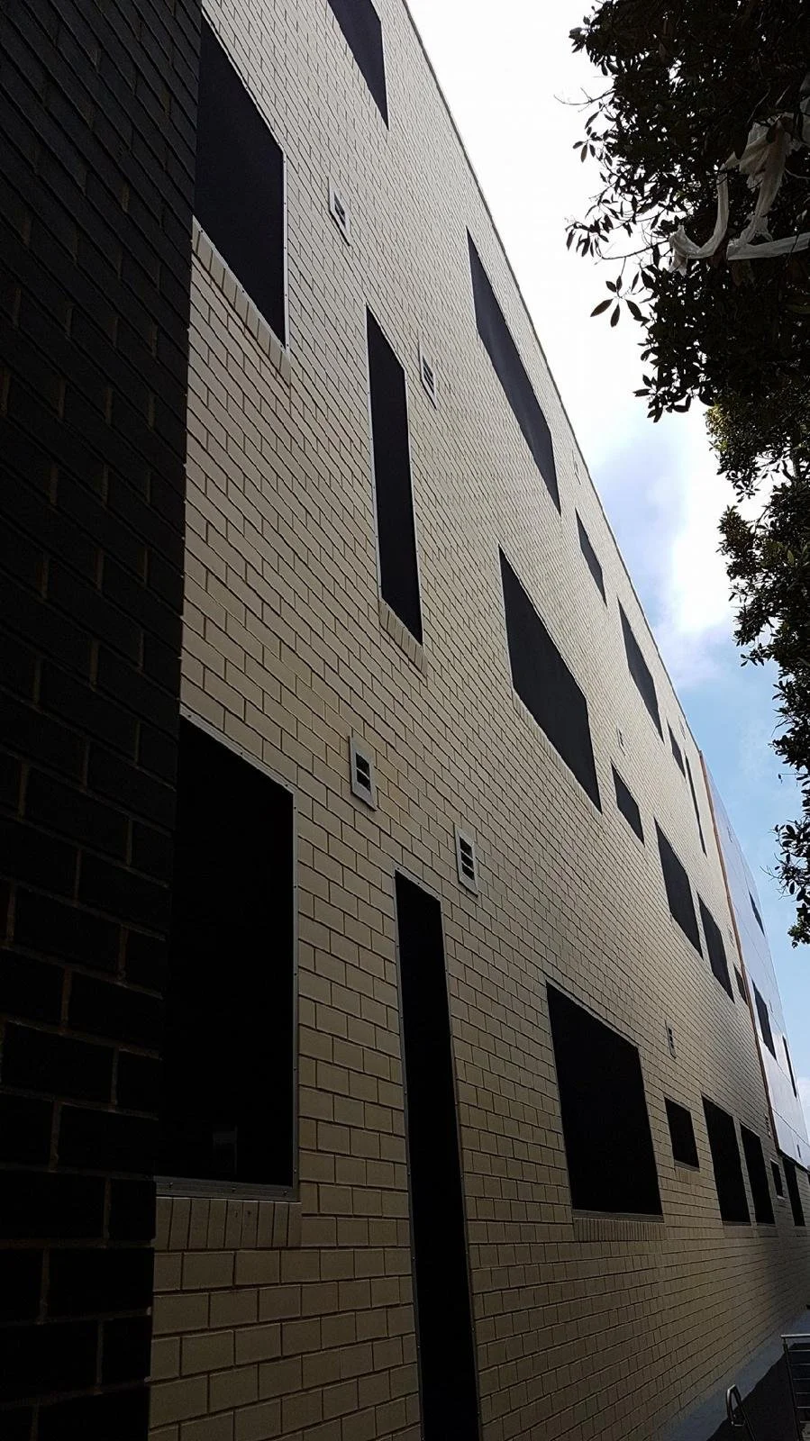 A multi-story building with beige brick exterior and several windows, some of which are covered or dark, seen from a low angle against a partly cloudy sky, with trees on the right side.