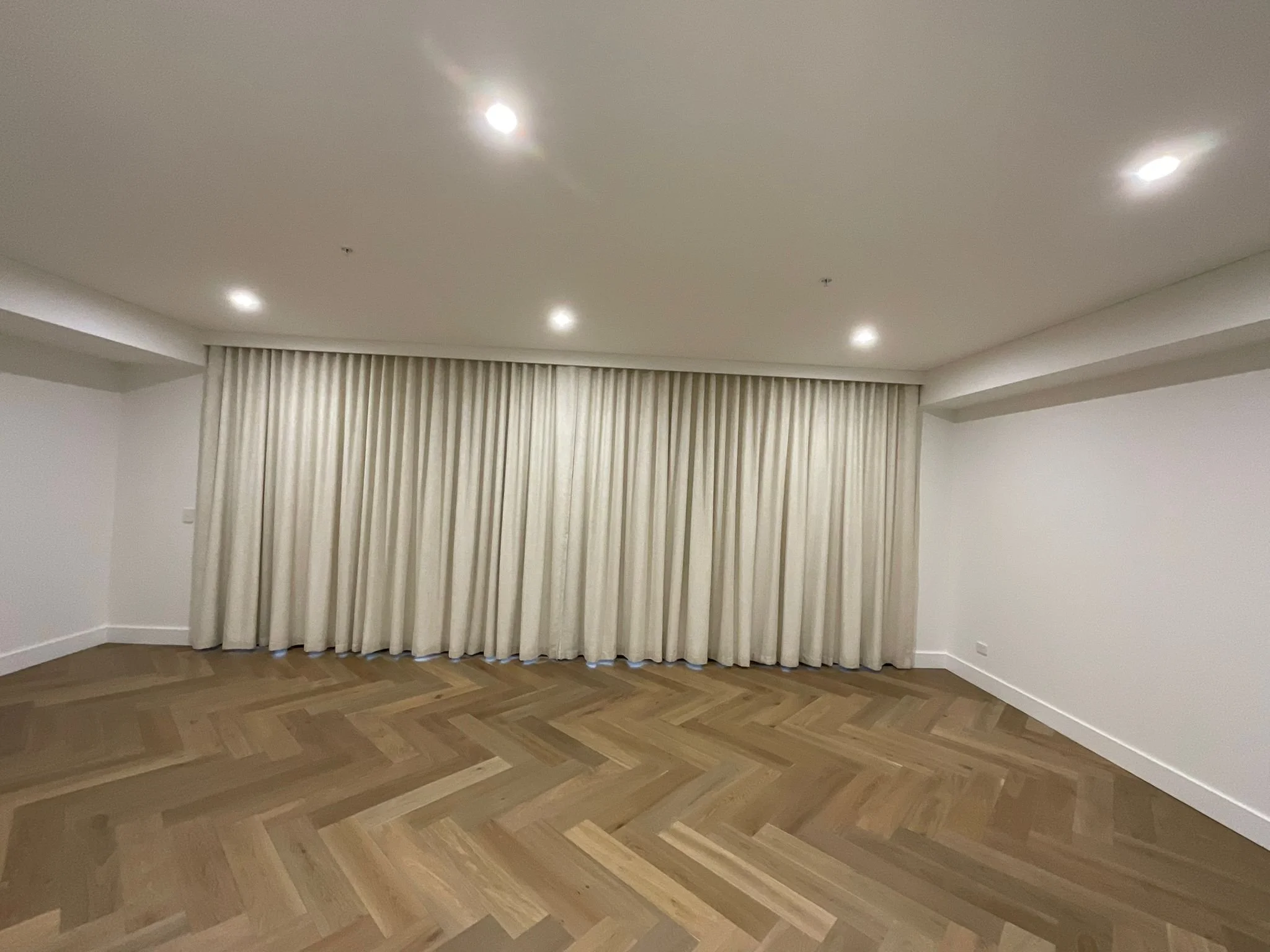 Empty room with wooden herringbone floor, beige curtains, white walls, and four ceiling lights.