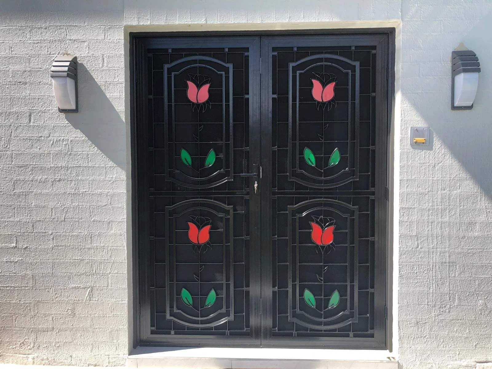 Black metal security door featuring red and green stained glass floral designs, mounted on a white brick wall with two wall lights on each side.