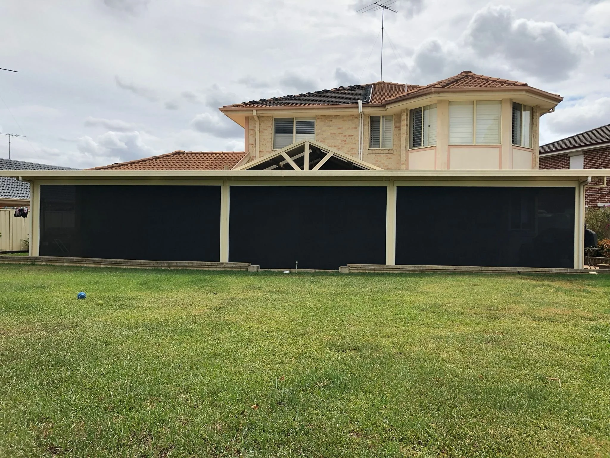 A backyard with a well-kept green lawn, scattered toys, and a large house featuring a brick facade, multiple windows, and a tiled roof. A black mesh fence surrounds part of the yard.