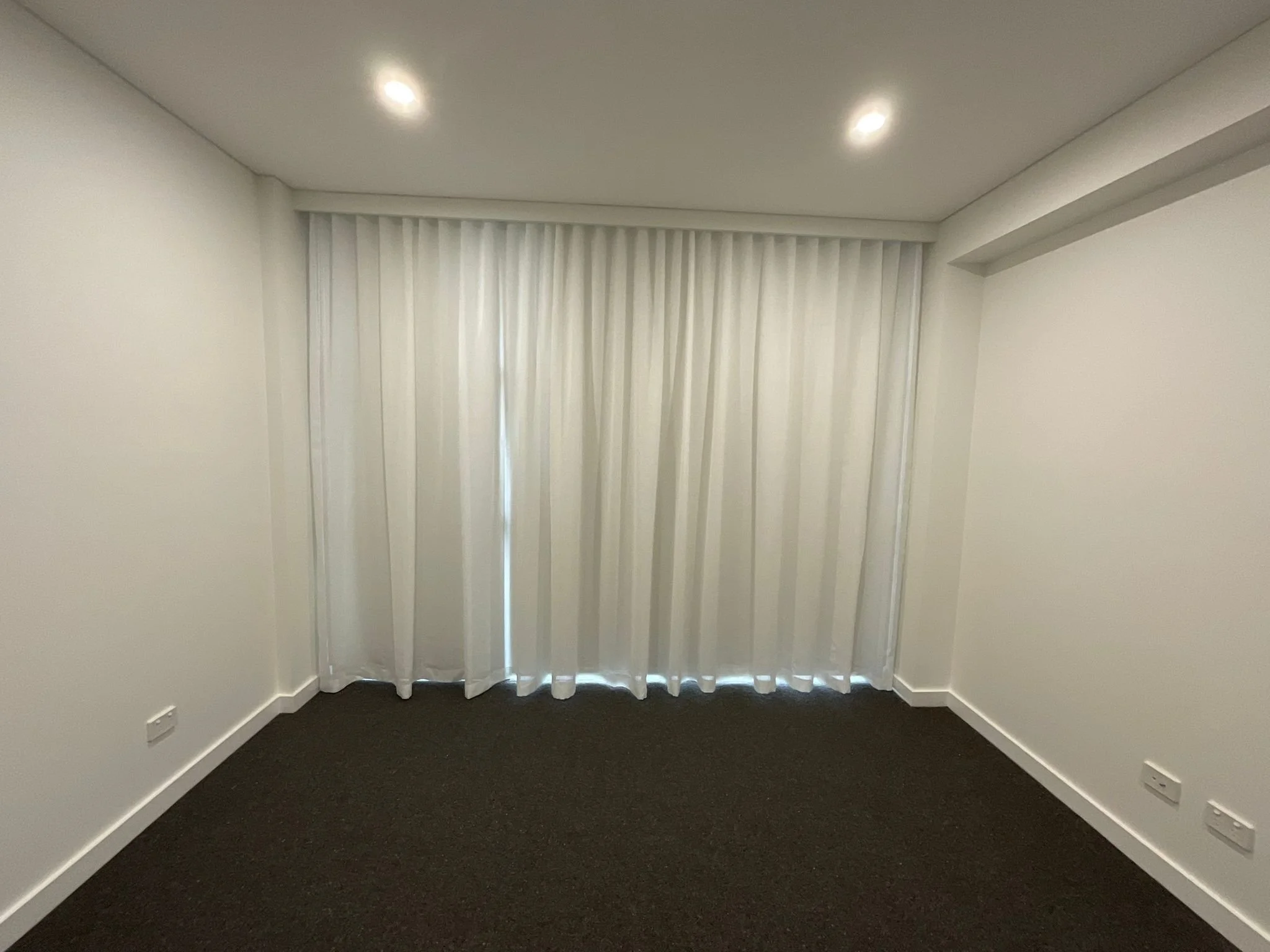 A room with white walls, a black carpet, and a large window covered by vertical blinds. The ceiling has two recessed lights.