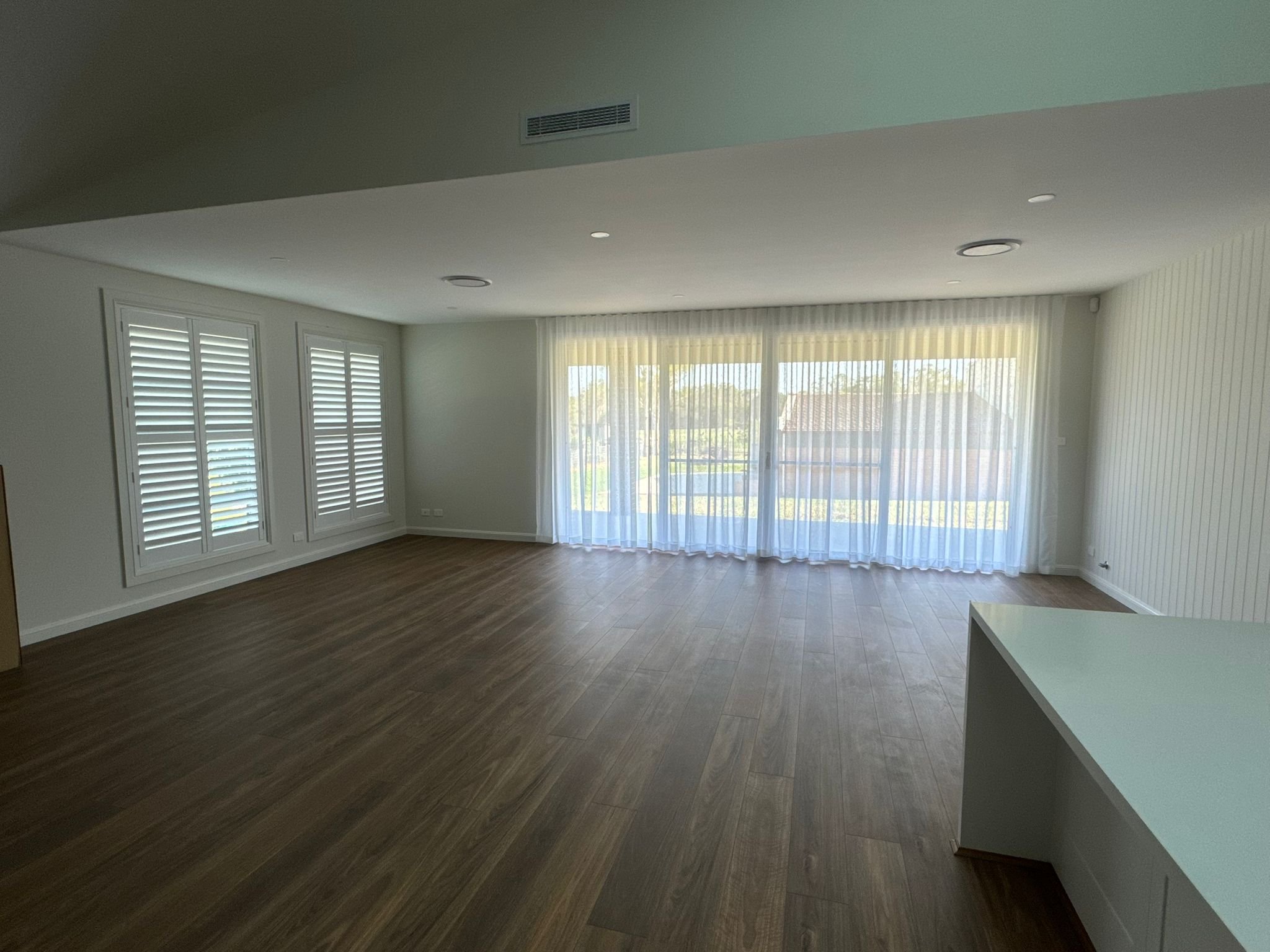 Empty living room with large windows covered with blinds and sheer curtains, hardwood flooring, white walls, and a section of a kitchen counter in the foreground.