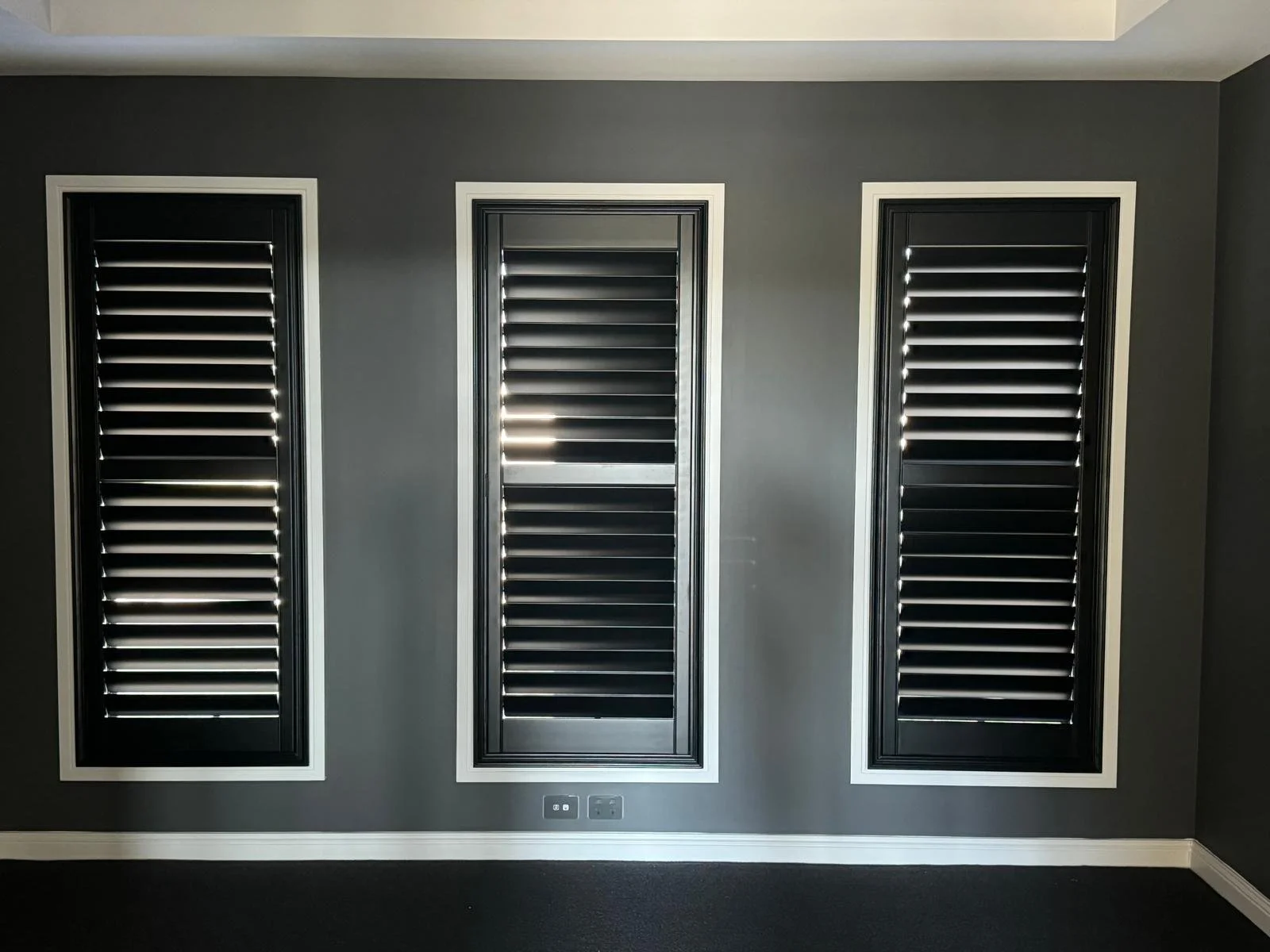 Three black window shutters with white borders mounted on a gray wall, with a black carpeted floor and electrical outlets below.
