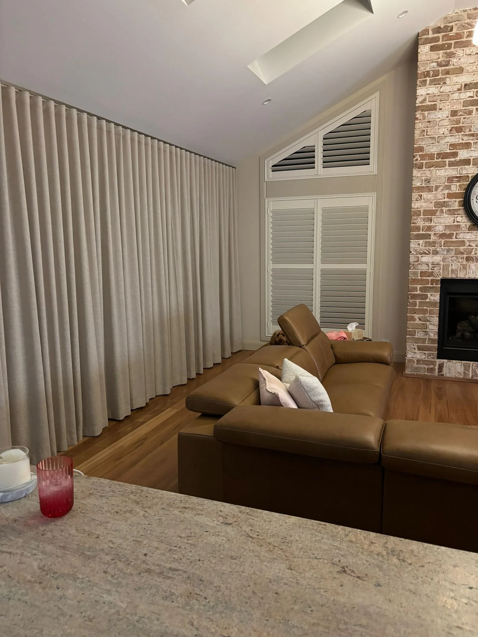 Living room with beige curtains, a brick fireplace, a tan leather sectional sofa, and a table with candles and a red vase.