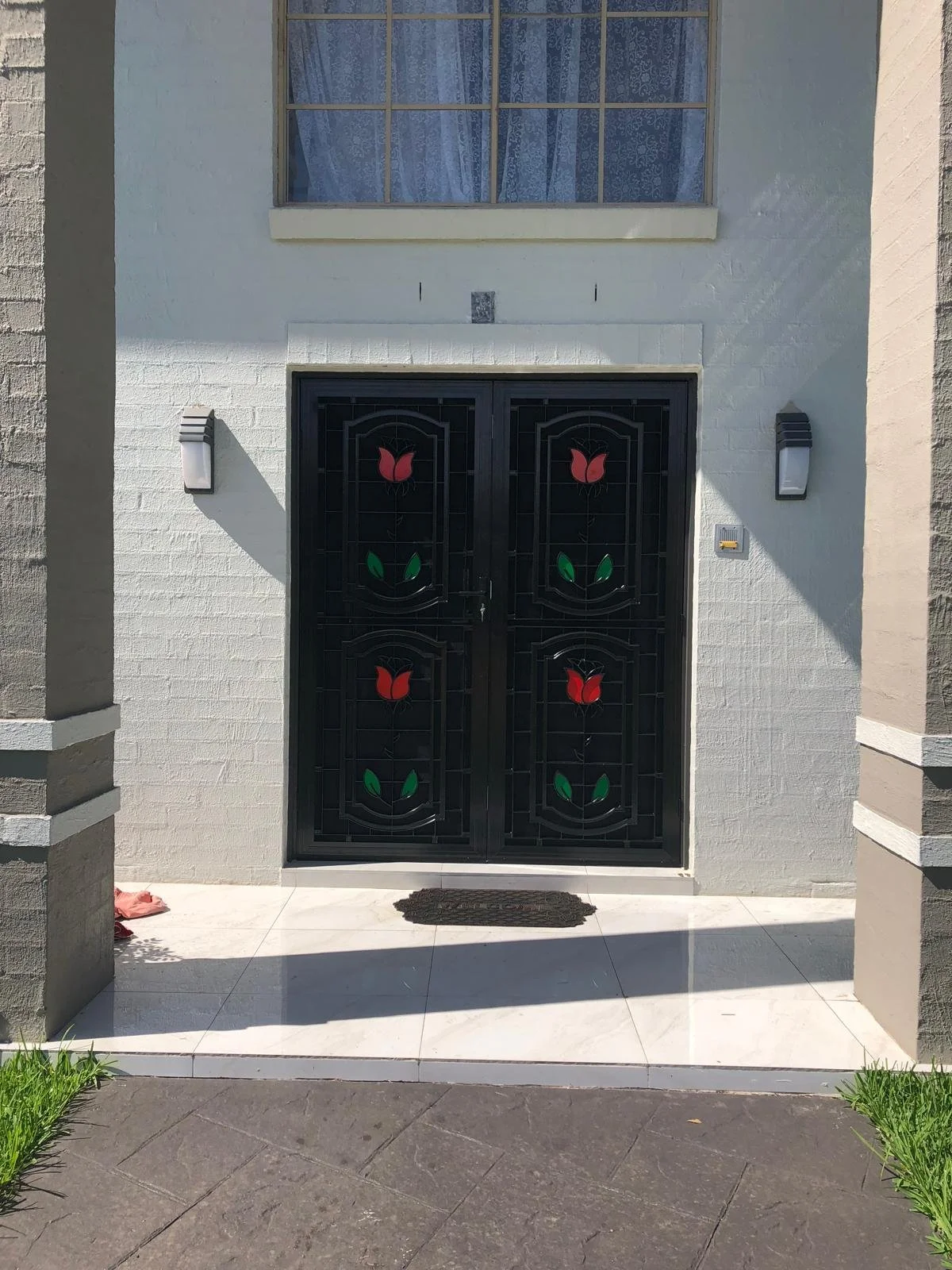 Front entrance with a black metal door featuring red tulip design and green leaves; wall-mounted lights on each side; white brick exterior with a window above and a small doormat.