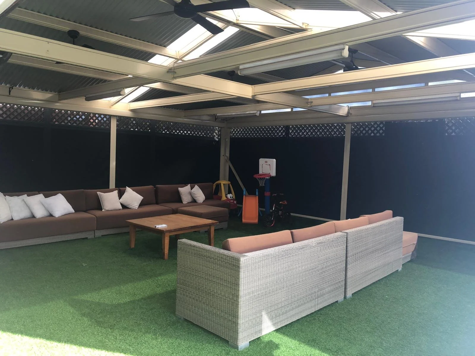 Outdoor patio with green artificial grass, a brown sectional sofa with white pillows, a wooden coffee table, beige woven sofa, and children's toys including a basketball hoop and a bicycle. The area is covered with a metal roof with skylights.