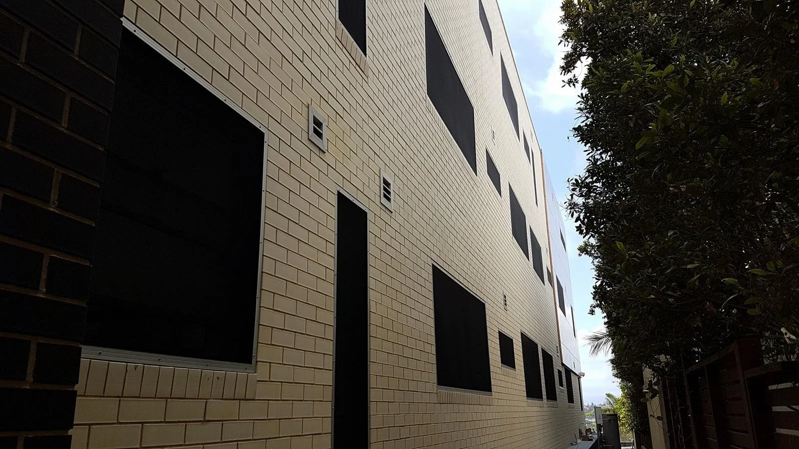 Side view of a modern multi-story building with beige brick exterior and multiple rectangular windows, some covered with black panels, along with a tree on the right side.