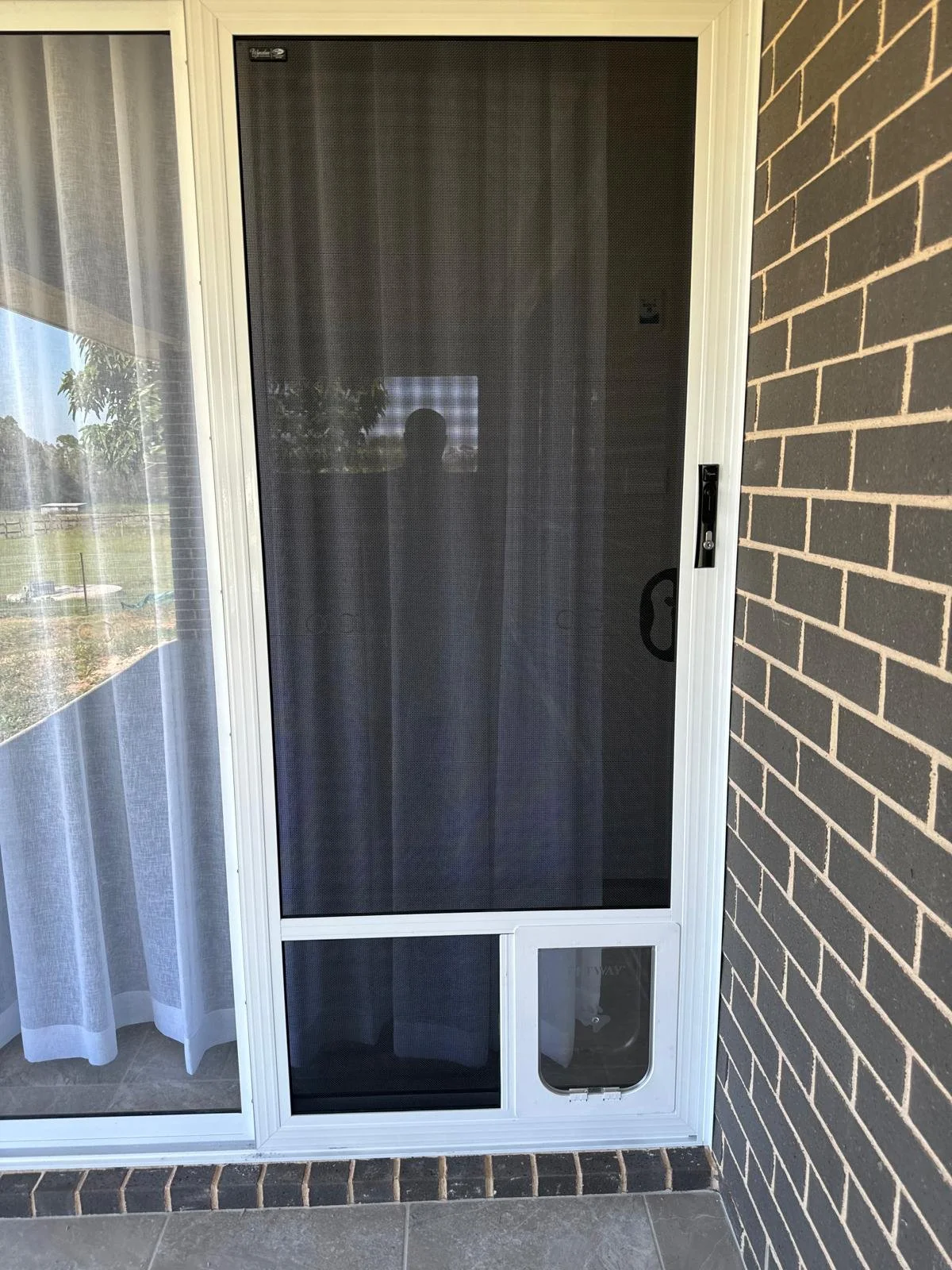 A glass door with a screen door in front, installed on a porch with a brick wall on the right and a glass window with curtains on the left. The screen door has a handle on the right side and a pet door in the bottom panel.