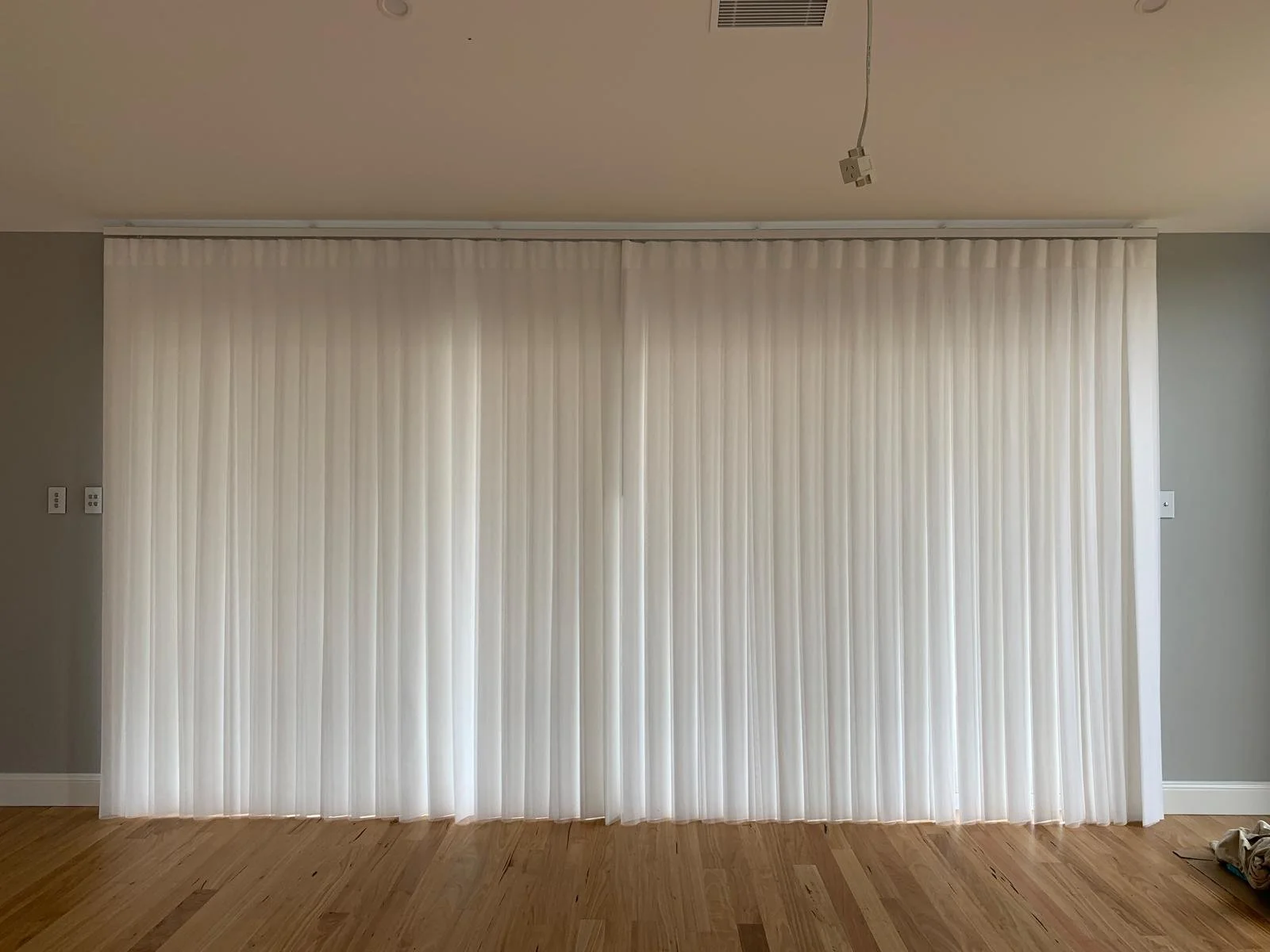 Closed white vertical blinds covering a large window or sliding door in a room with gray walls and a wooden floor.
