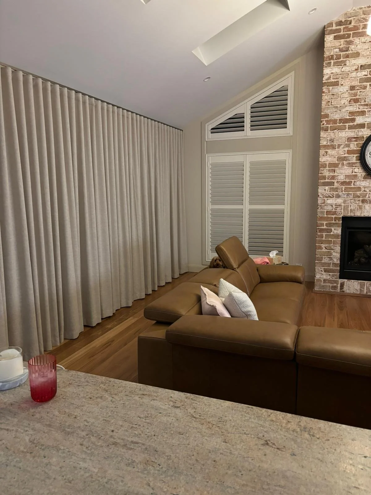 Living room with beige curtains, brown leather sofa with pillows, brick fireplace, wall clock, and a small dog peeking from behind the sofa.