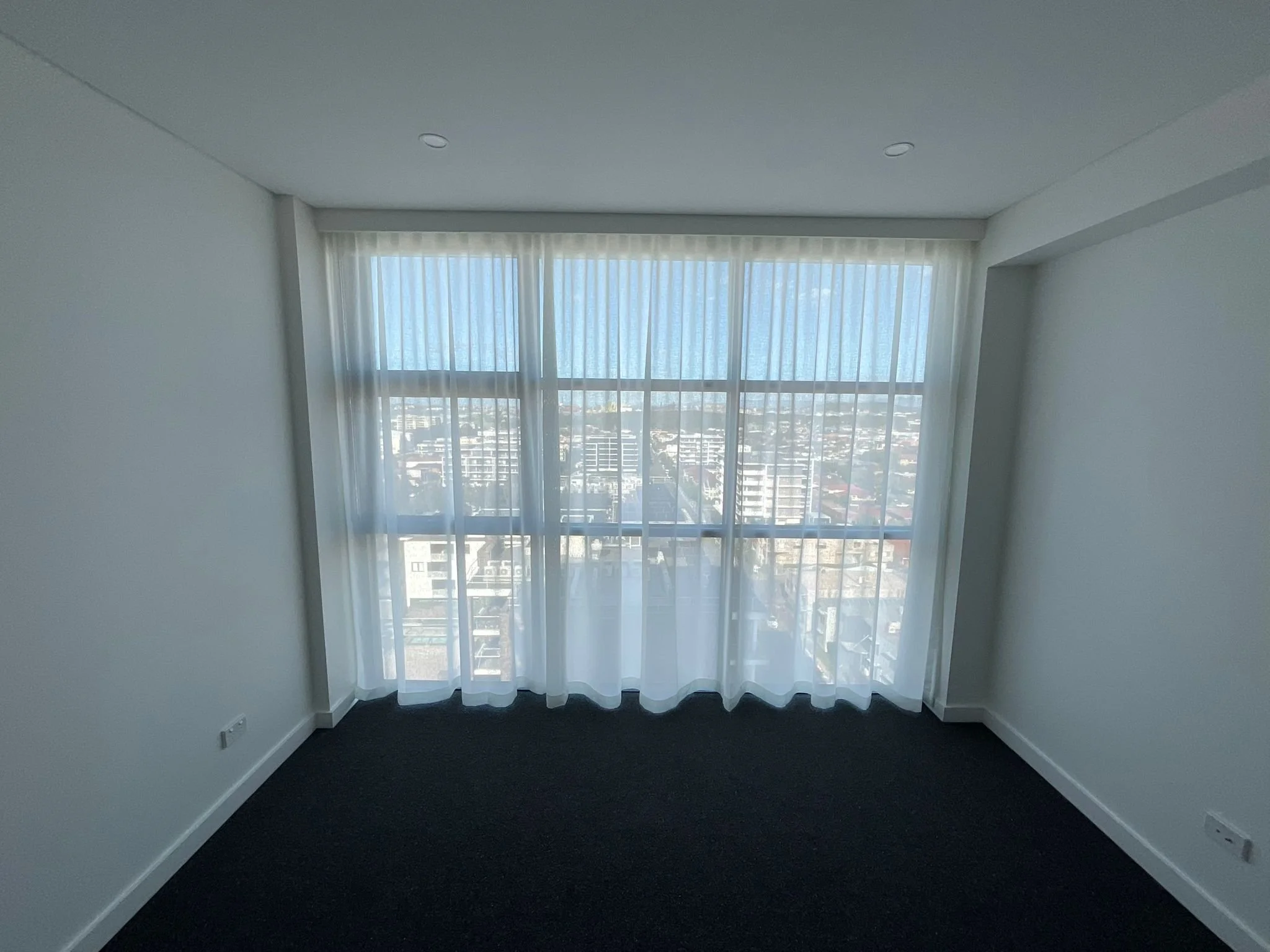 A room with large windows covered by white curtains, view of city buildings outside, dark carpeted floor, white walls, and ceiling with recessed lighting.