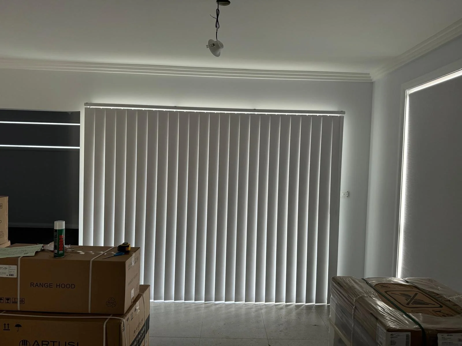 Interior room with closed vertical blinds covering large windows, construction boxes, and a ceiling light fixture without a bulb.