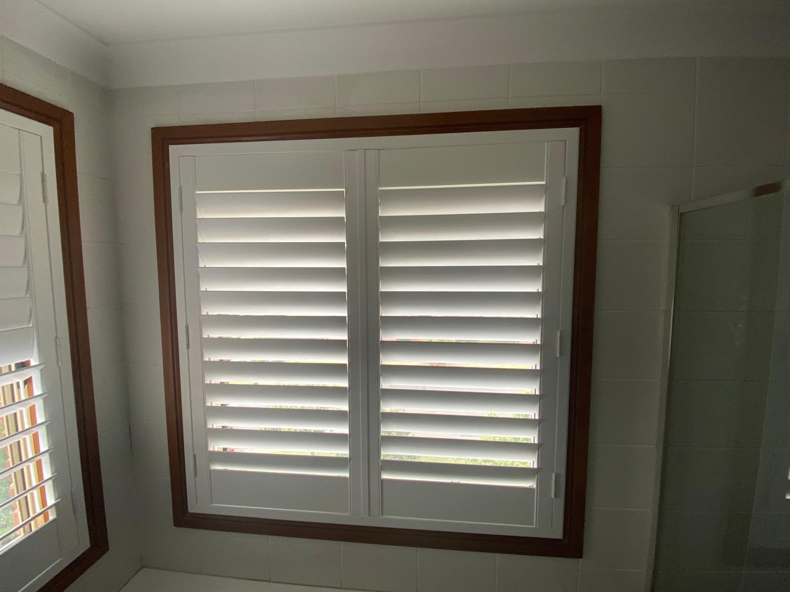 Interior view of a room with a large window featuring white blinds framed by wood, a small window with blinds, and tiled walls.