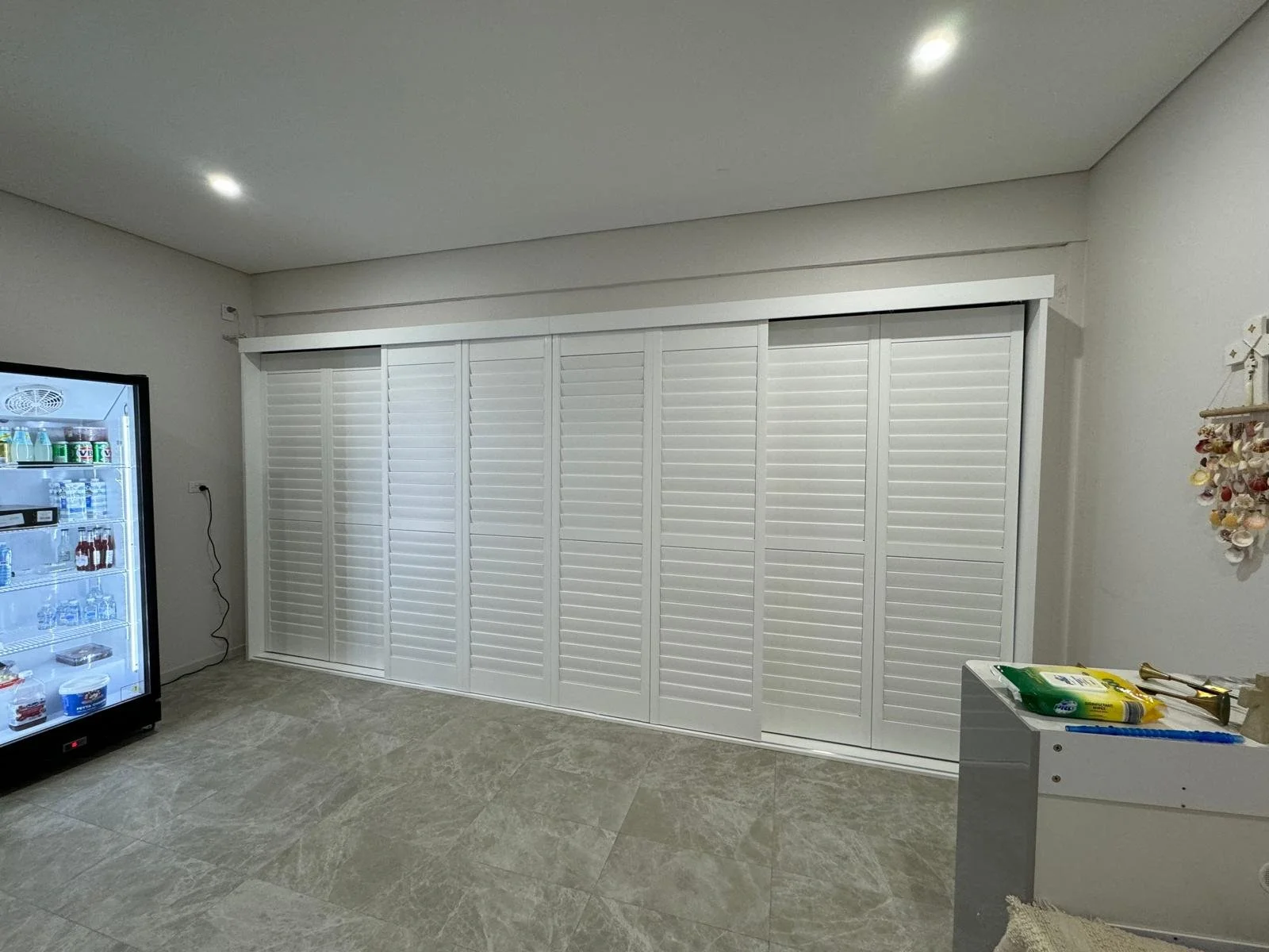 Room with white sliding closet doors, a beverage refrigerator on the left, and some items on a small table on the right, including a pack of wipes and a brass object, with shell decorations on the wall to the right.