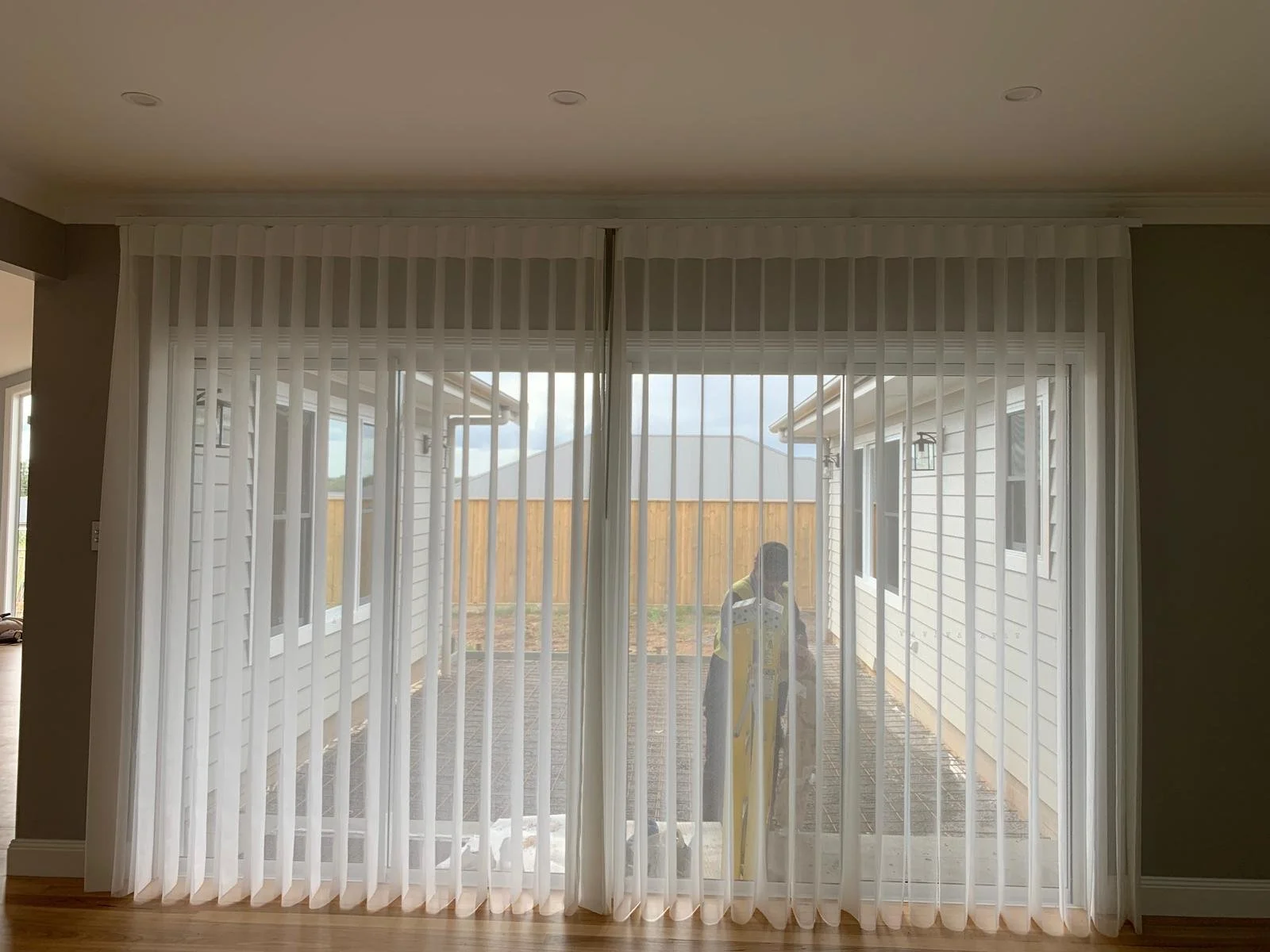 View through a sliding glass door with vertical blinds, showing a person outside near a house with white siding and a wooden fence in the background.