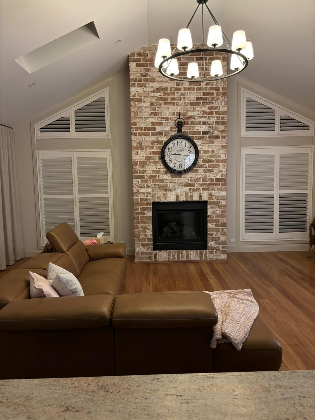 Living room with brick fireplace, large clock above the fireplace, brown leather sectional sofa with pillows, wooden flooring, and white window shutters. A chandelier hangs from the ceiling.
