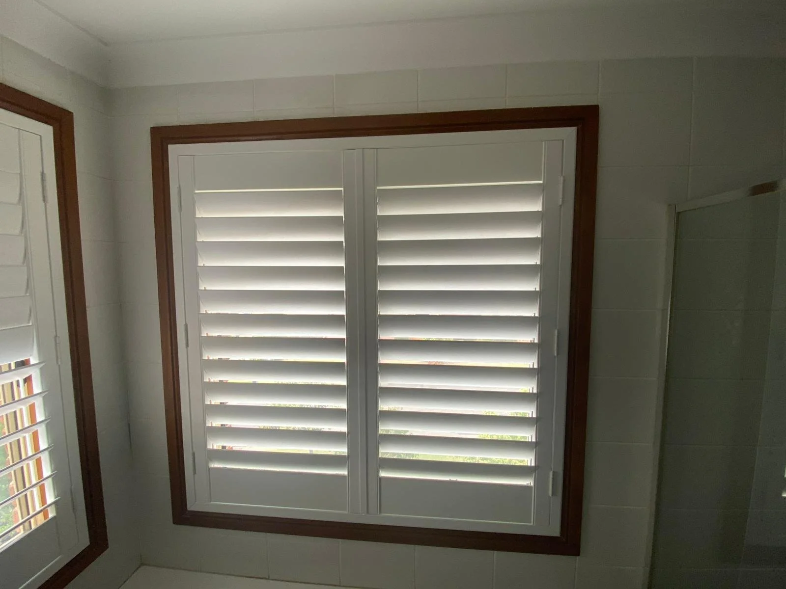Bathroom with window featuring white plantation shutters, brown trim, and beige tiled walls.
