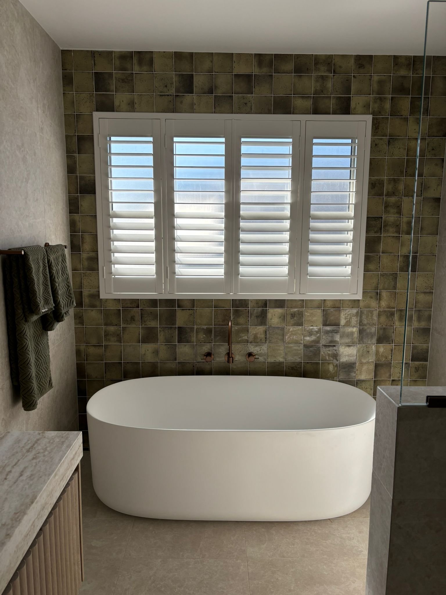 Modern bathroom with a freestanding bathtub, a large window with white shutters, and tiled walls and floor.
