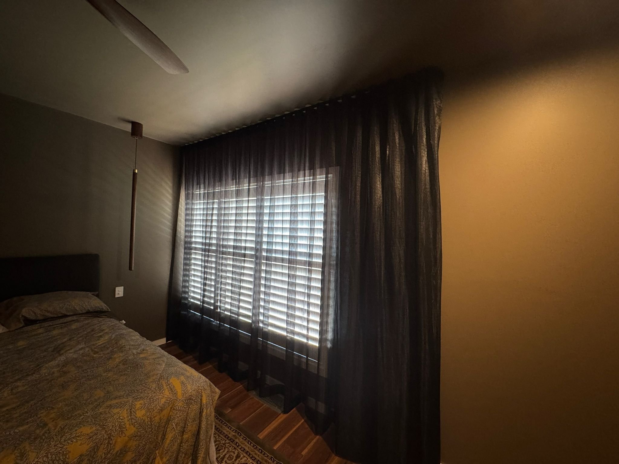 A bedroom with a bed in the left corner, partially visible, with a patterned bedspread. Next to the bed is a wooden lamp with a fabric shade. The room has a large window with partially closed blinds and sheer dark curtains. The wall behind the window