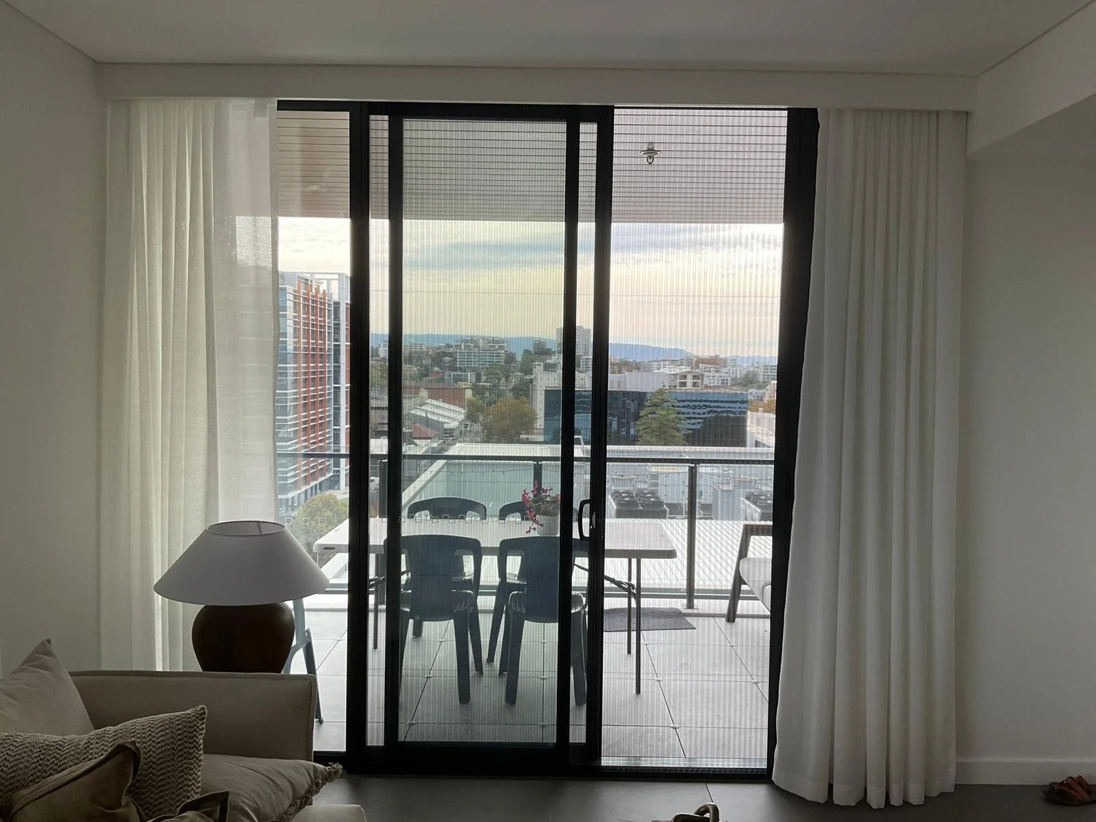 Living room facing a balcony with a view of a cityscape and distant mountains, sliding glass door, white curtains, a table with four chairs, a potted plant, and a lamp.