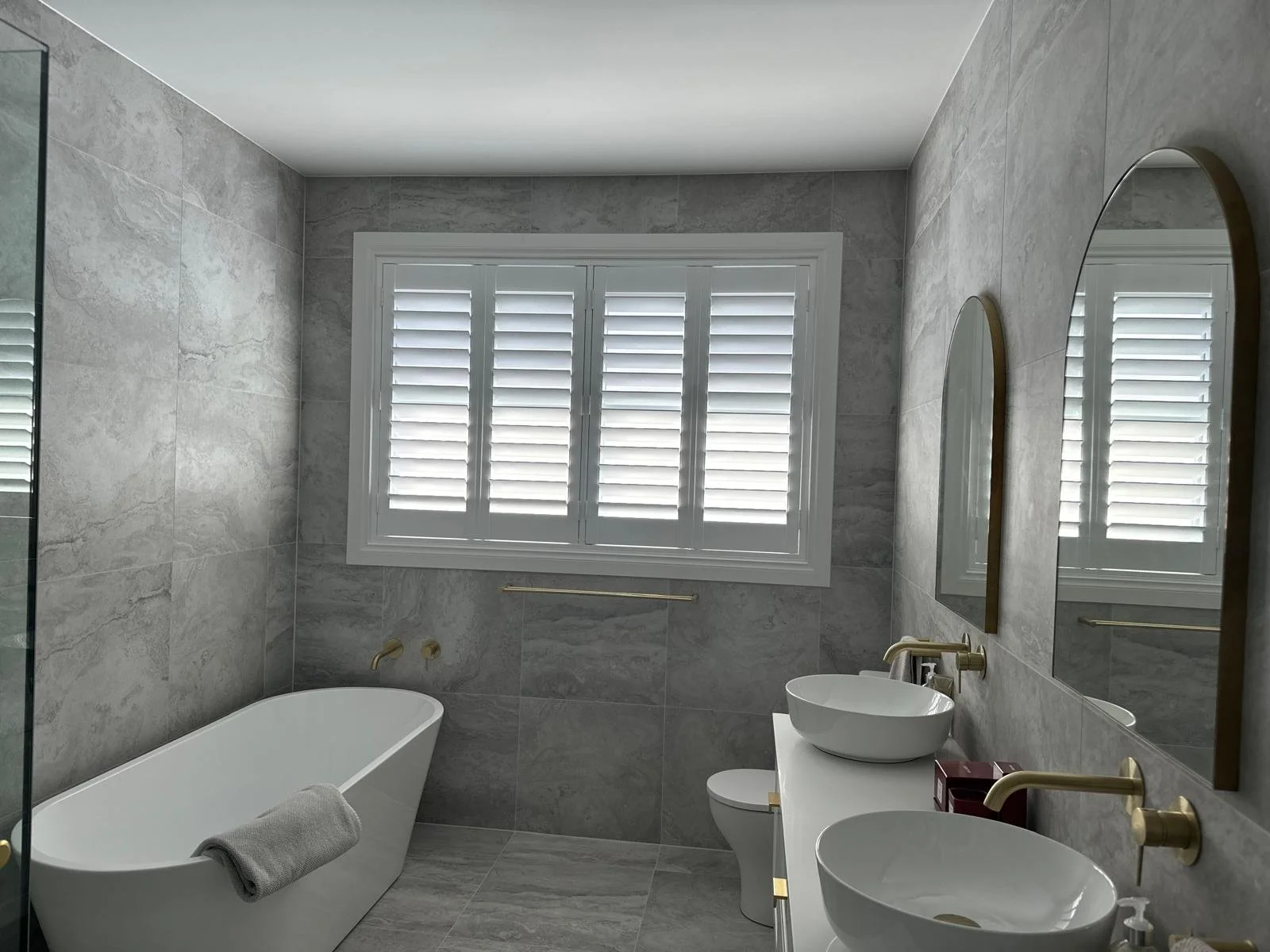 Modern bathroom with gray marble walls, a white bathtub, a window with white shutters, and two white vessel sinks with gold fixtures.