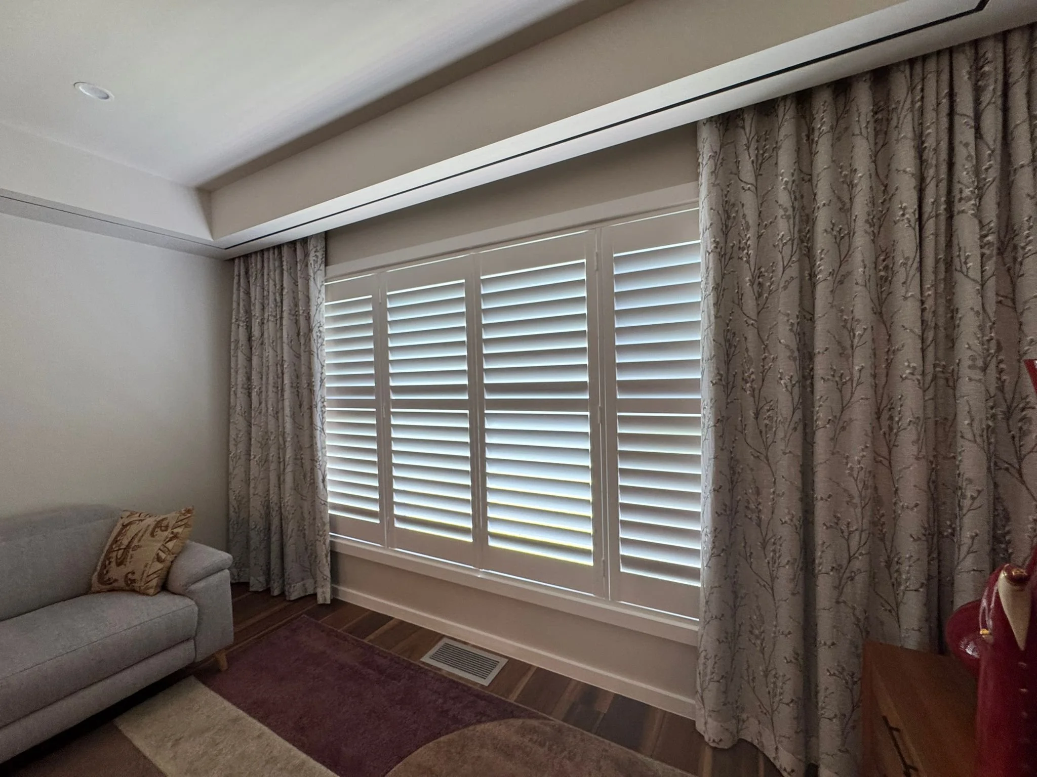 A living room with a large window covered by closed white plantation shutters and floral curtains on each side. Part of a gray sofa with decorative pillows is visible in the lower left corner. The floor has dark wood planks and a dark rug. There is a