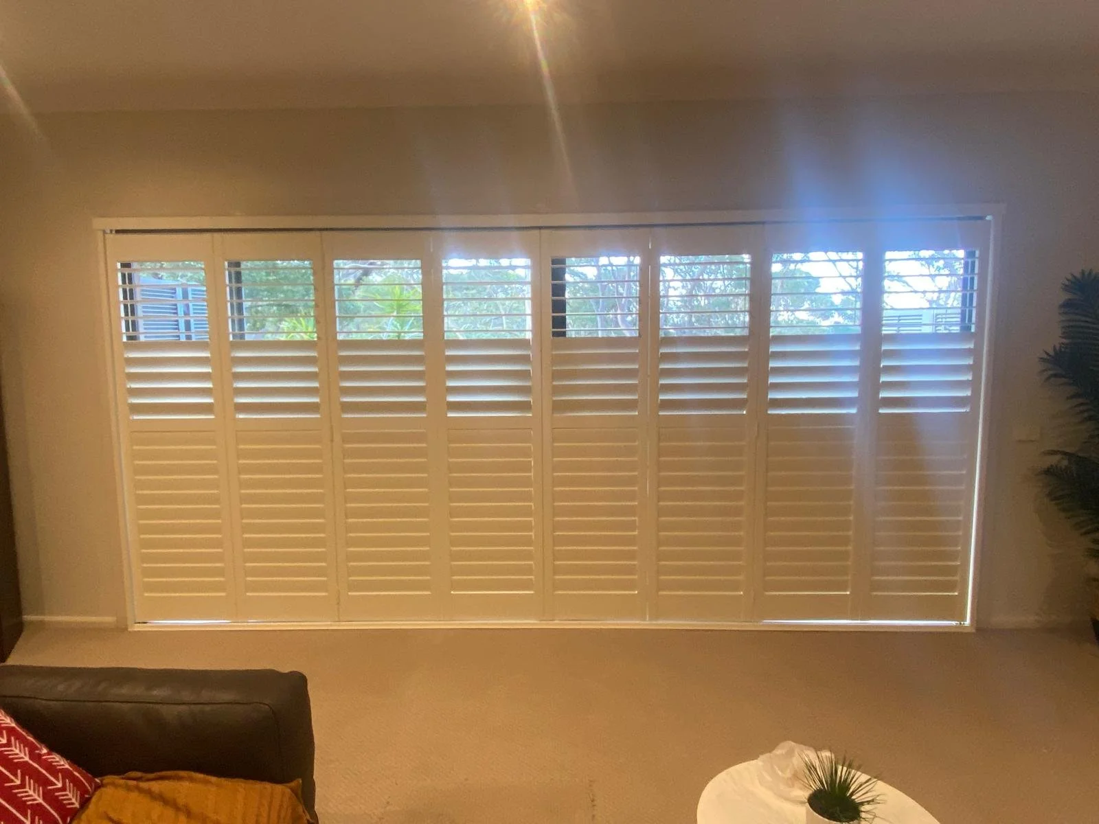 Living room with large window covered by white plantation shutters, showing green trees outside, with a brown leather sofa and a small round white table with a plant inside.