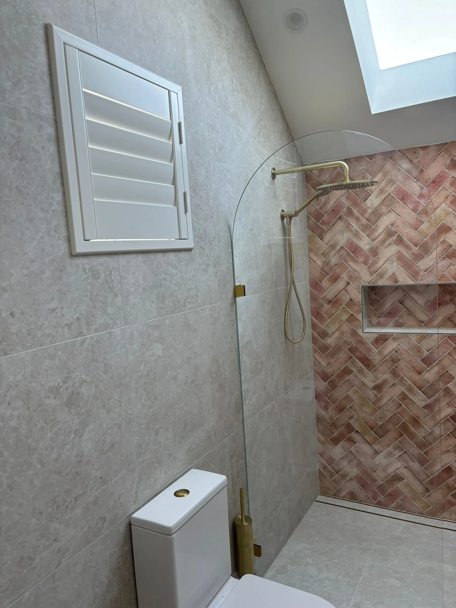 Modern bathroom with light gray tiled walls, a white toilet with a gold flush button, a chrome showerhead, and a glass shower partition. There is a skylight window on the ceiling illuminating the space.