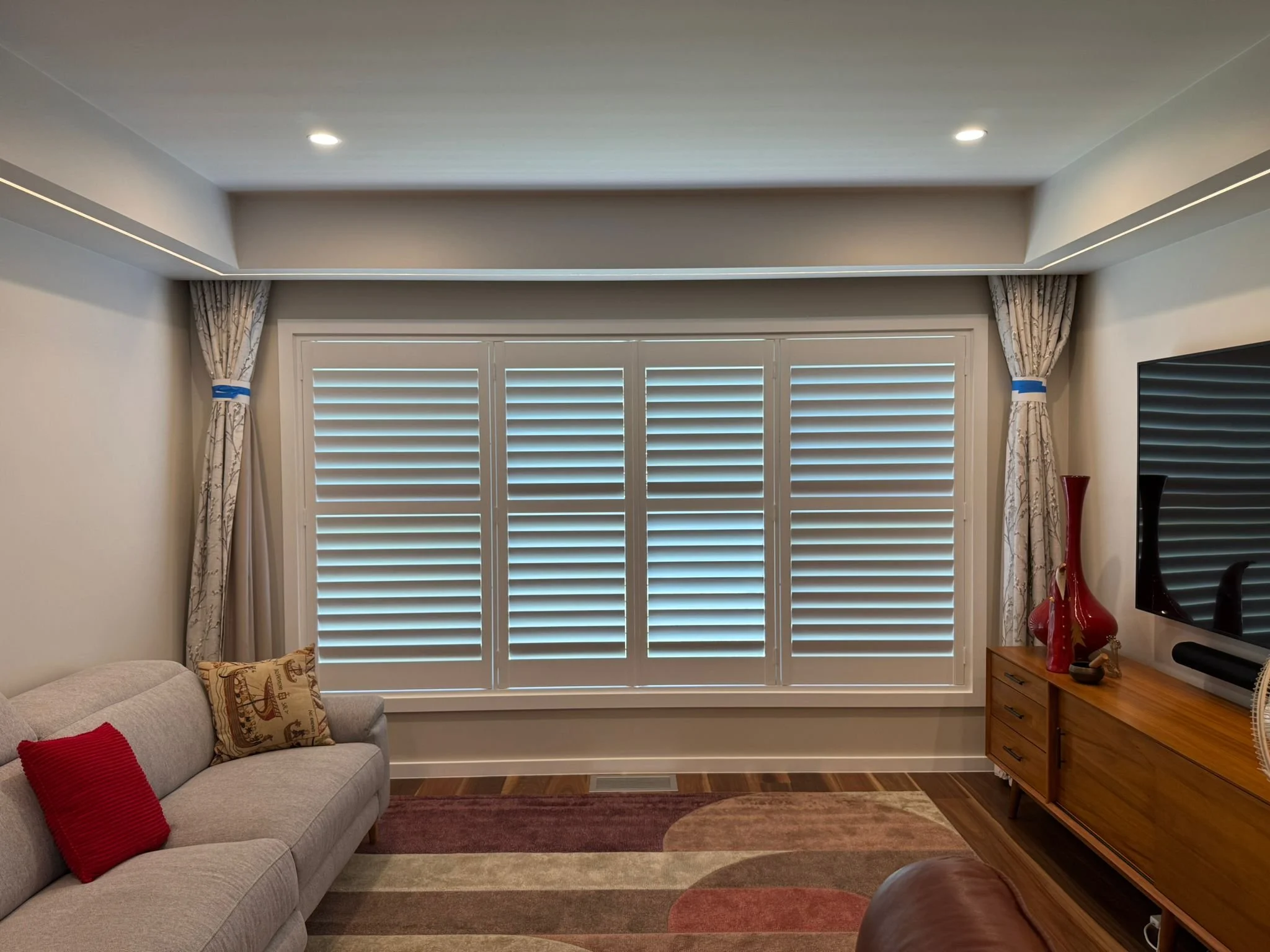 Living room with a large window covered by white plantation shutters, beige walls, a beige sofa with patterned cushions and red accent pillow, a wooden dresser with decorative vases, and a television mounted on the wall.